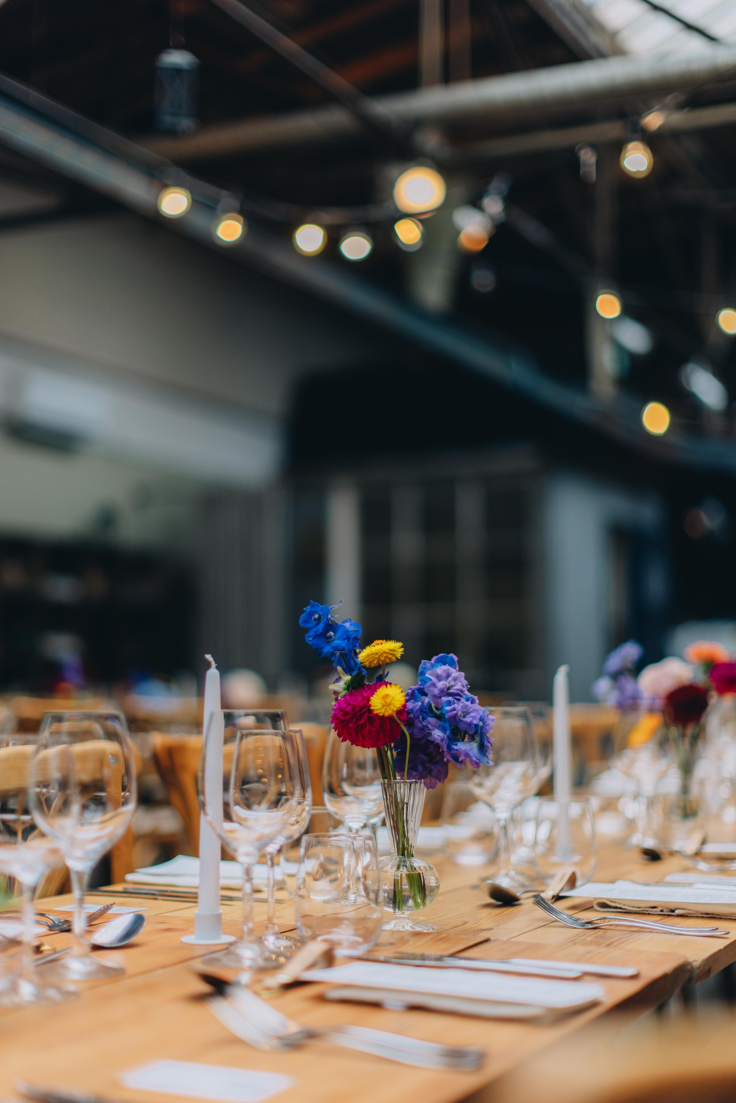 Bord med farverige blomsterdekorationer, tomme vin- og vandglas, tallerkener, bestik, lysestager med hvide lys og tekstiler, inde i en indretning med bløde belysning og loftstrukturer.
