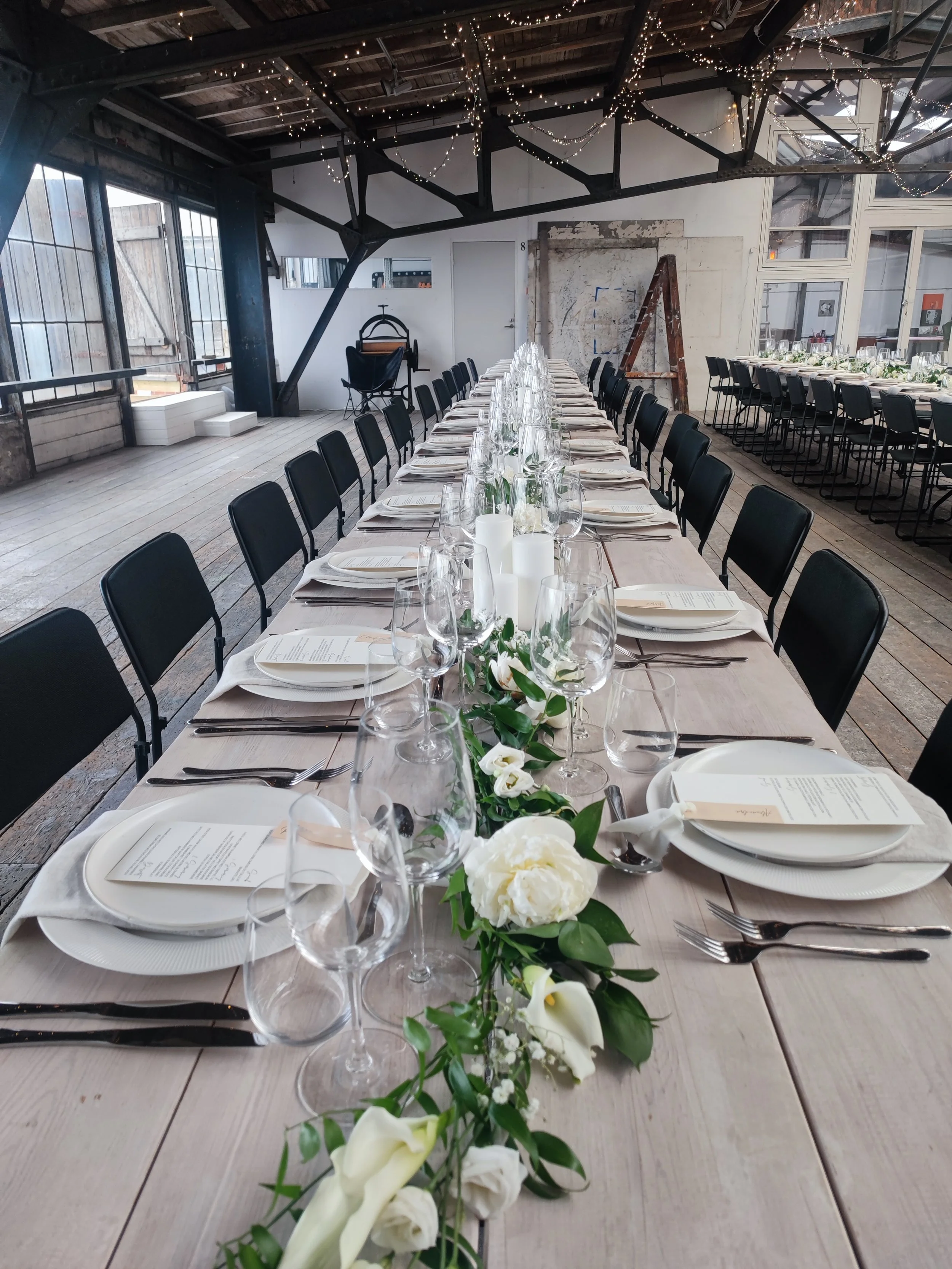 Langbord dækket til fest med hvide tallerkener, bestik, vin- og vandglas, papirmenu og hvid blomsterdekoration med grønne blade.