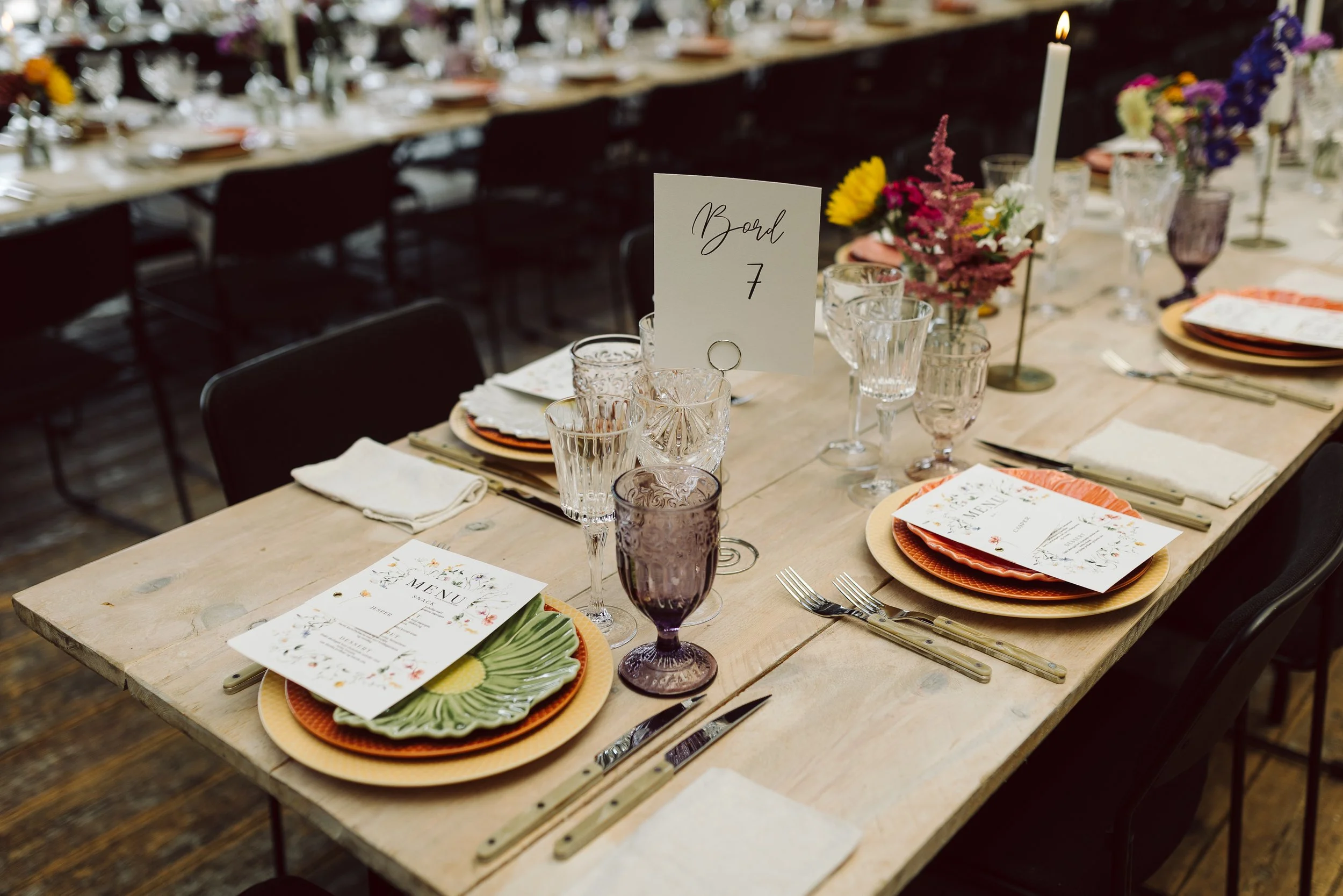 Bordopsætning med tallerkener, glas, bestik, bordkort og blomsterdekorationer til en festlig begivenhed.