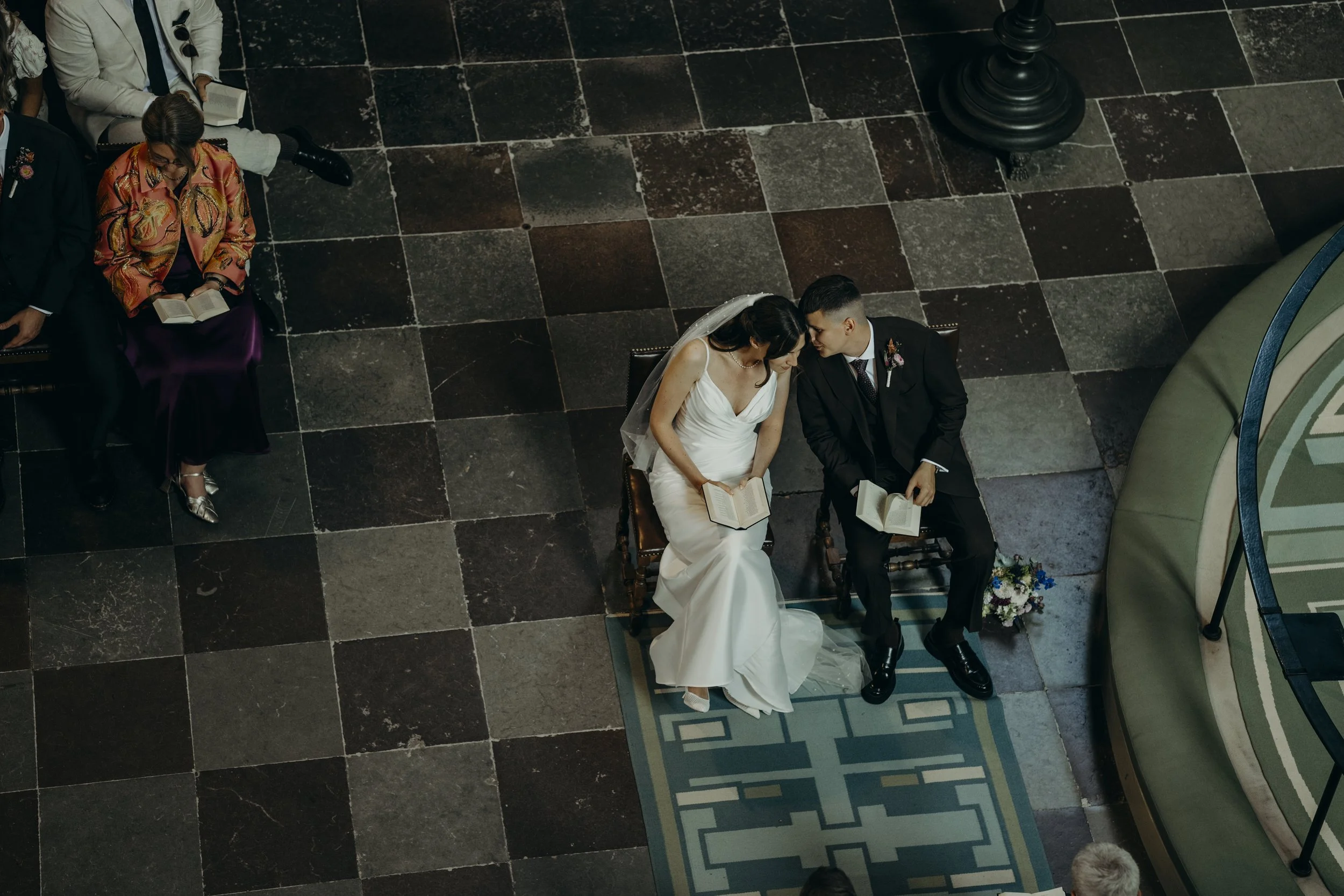 En bryllupsceremoni med en brud og en gommepå en bænk, der holder hinanden i hænderne og ser på hinanden, alle med bøger i hånden, set fra oven.