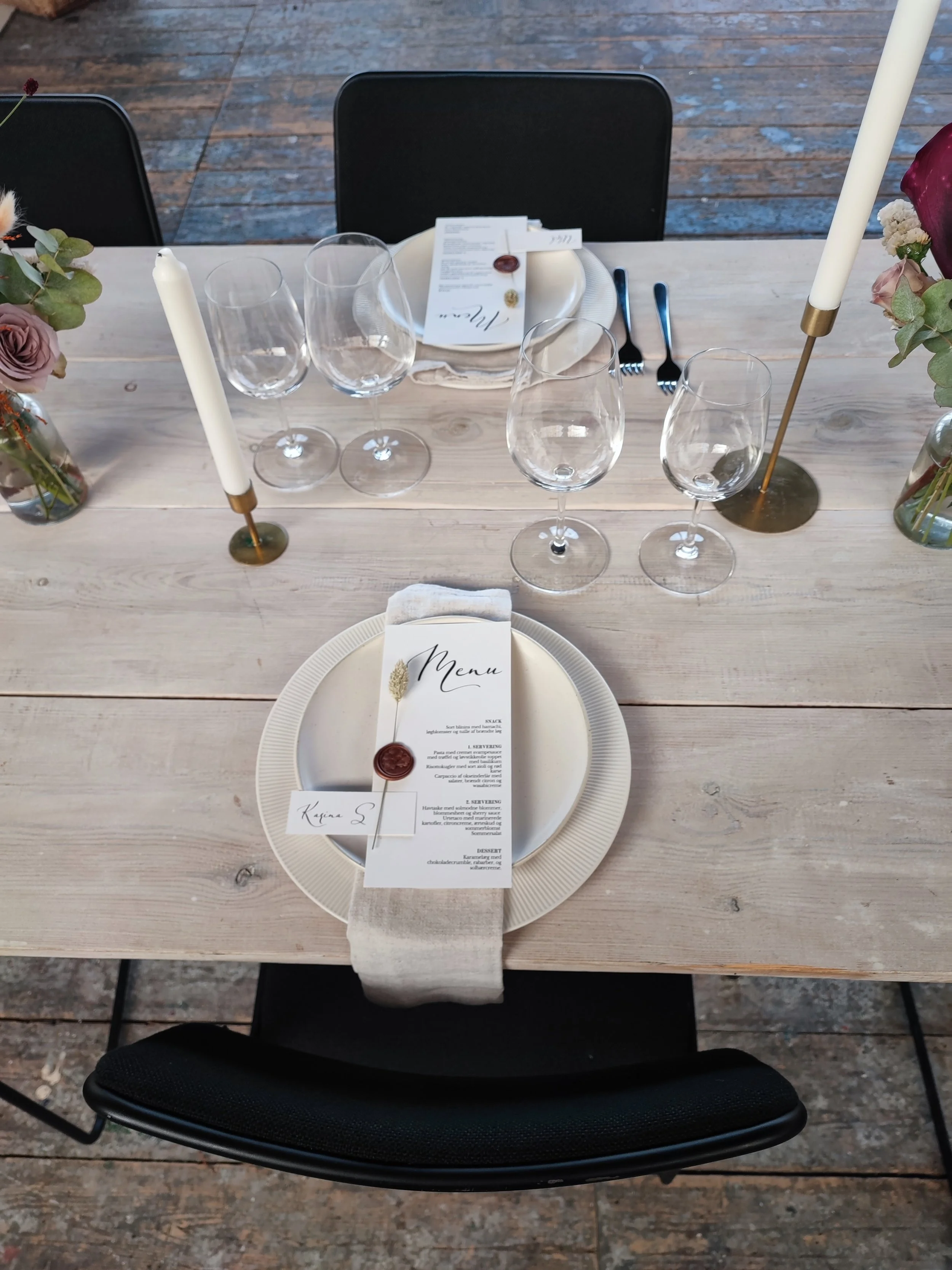 Borddækning med et rundt fad med menu, tre vinglas, tallerken med serviet og menu, stearinlys og blomster.