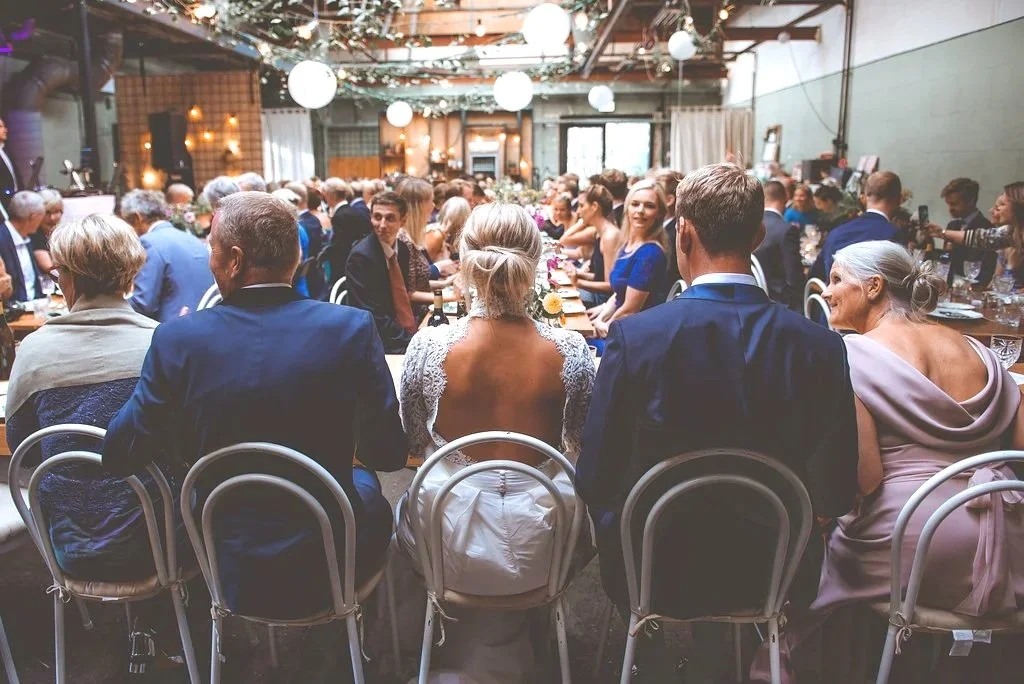 En gruppe mennesker sidder ved langbord i et festlokale, sandsynligvis til en bryllupsfest, med pynt og lamper hængende fra loftet.