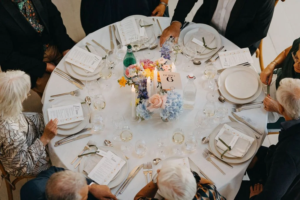 Middagsselskab med otte personer siddende omkring et rundt bord dækket med bordservice, glas, bestik, menukort og en blomsterdekoration i midten.