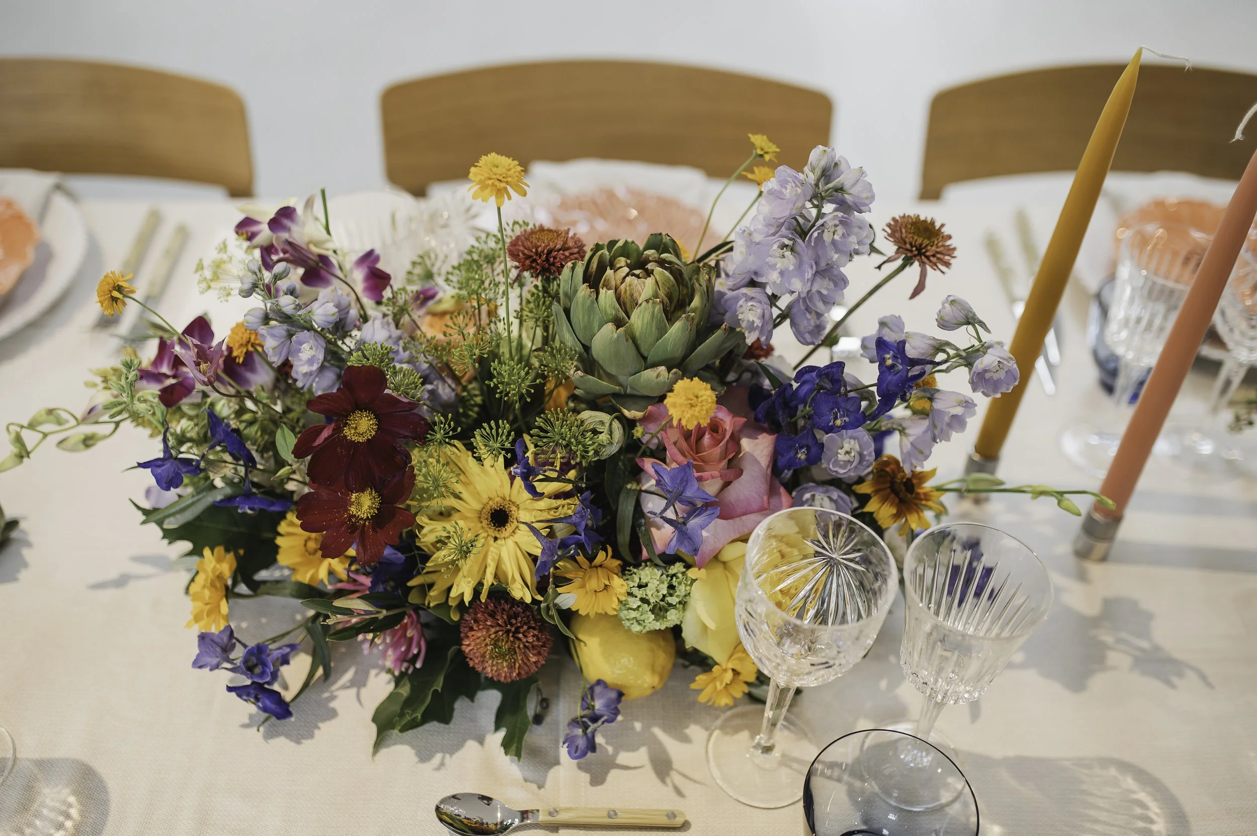 Farvet bryllup blomsterarrangement af Bliss Cph med roser, protea, delphiniums og zinniaer på bord med italienske krystalglas glas og  farverige stearinlys.