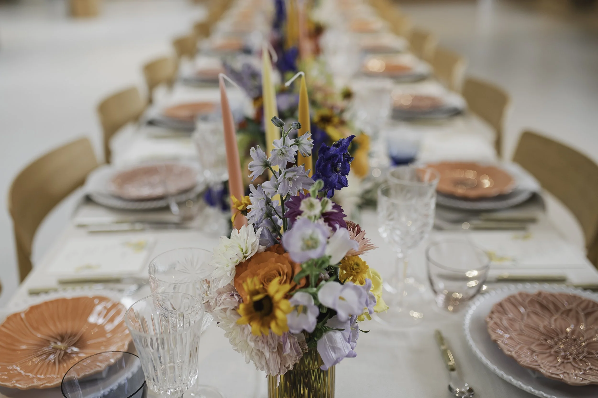 Borddækning med en lang borddekoration af farverige blomster og stearinlys, med fine glas og serveringsservice til en festlig begivenhed af Bliss Cph.