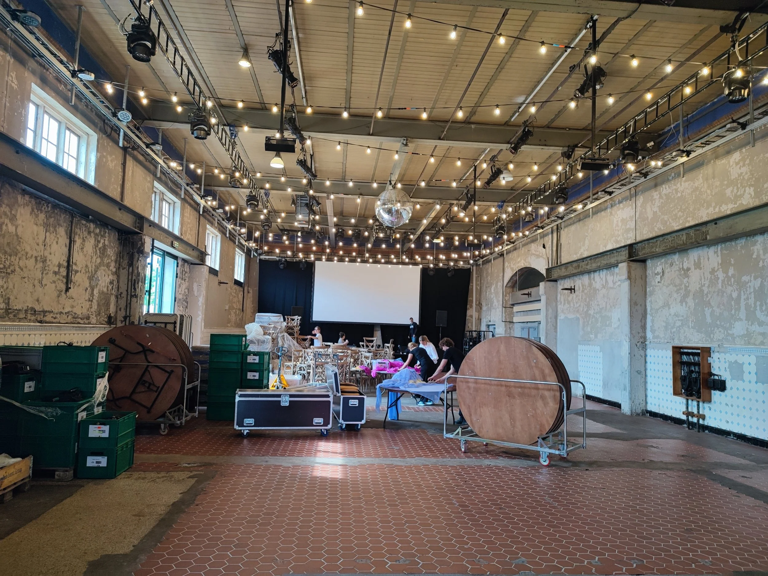 Indendørs koncert- eller eventlokale med gamle vægge, højt til loftet, dekoreret med mange strategisk placerede lysrør. Der er et stort projektor- eller lærred på den forreste væg. Personer arbejder med opstilling af udstyr, stole og borde foran scenen.