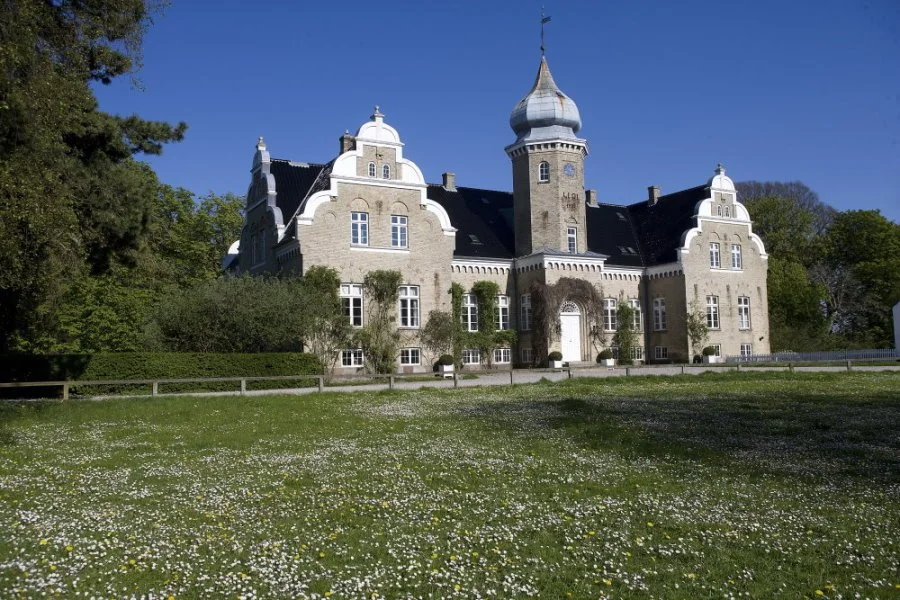 Bliss Cph - Søllestedgård Gods, en stor historisk bygning med tårne, omgivet af grønne træer og en blomstereng foran.