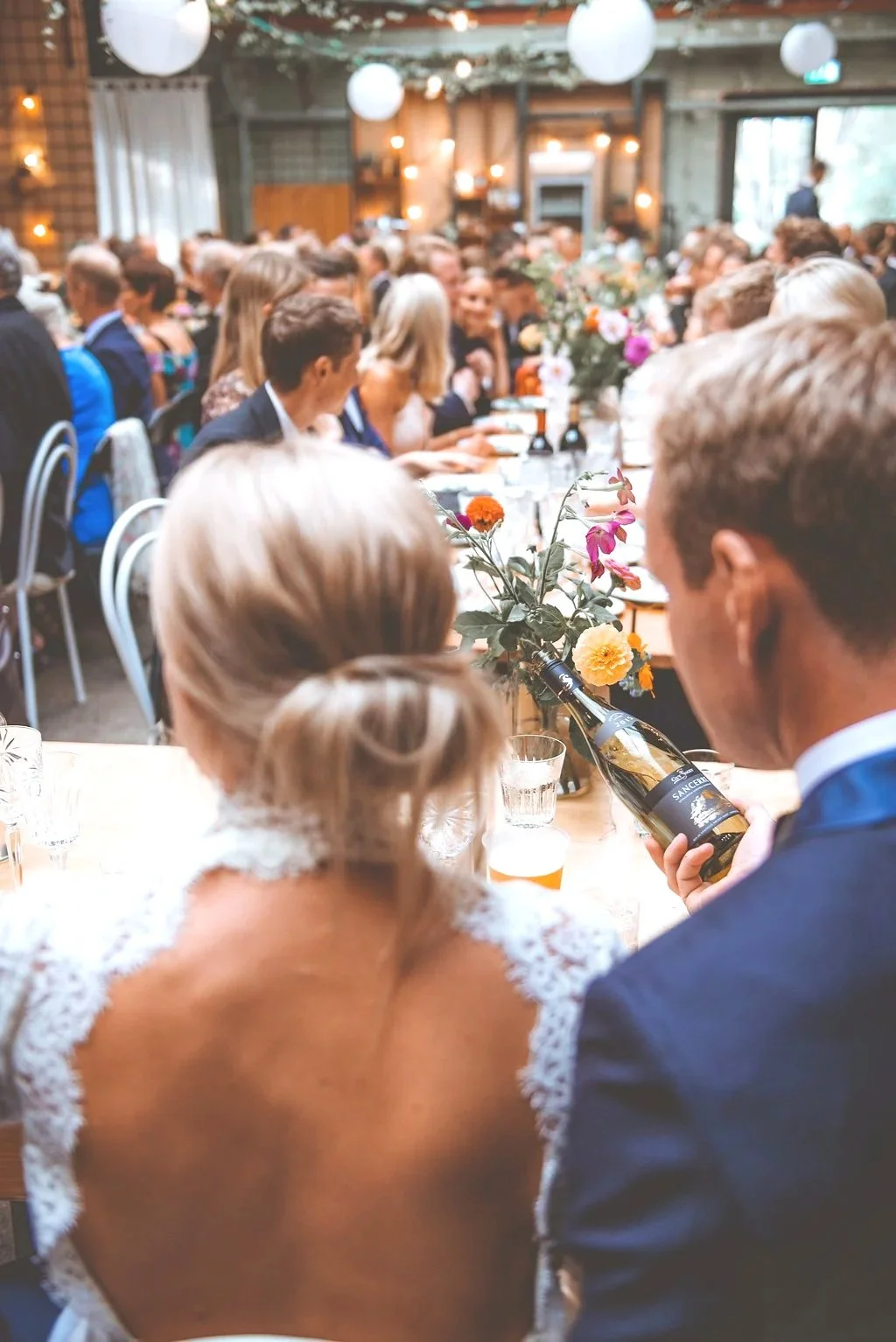 Et festligt arrangement hvor folk sidder ved langbord dekoreret med blomster, og en mand giver en flaske vin til en kvinde. Mange personer i festtøj, og lyse lys og paperlanterns pynter lokalet.