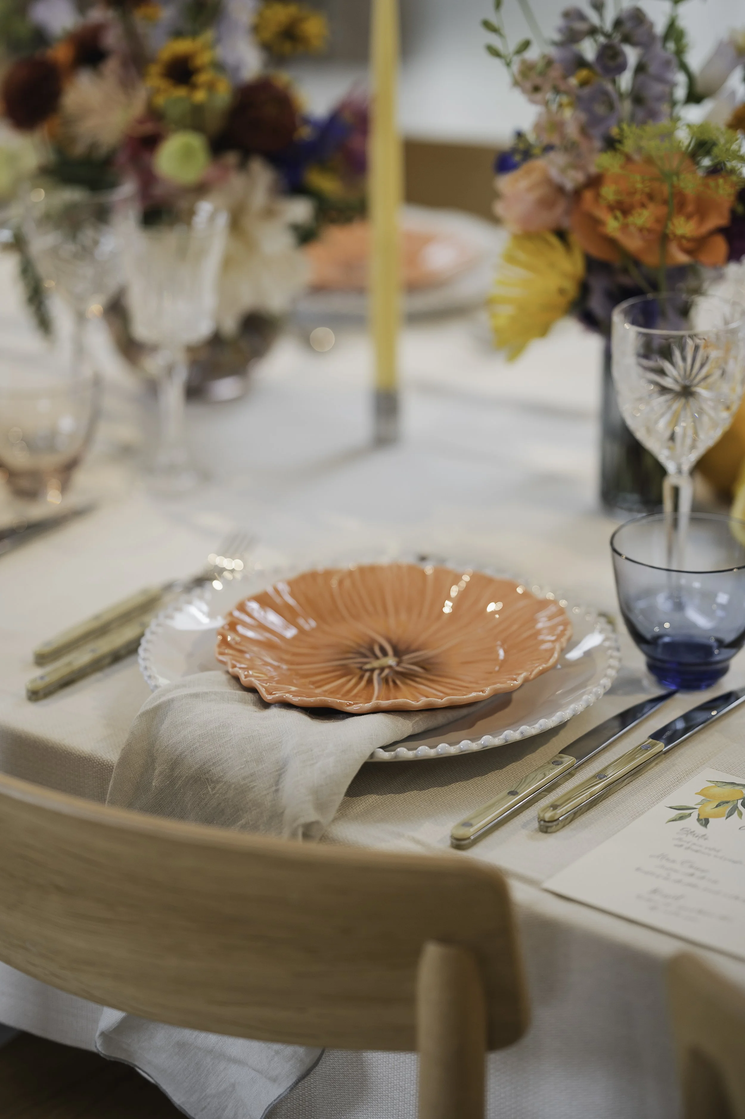 Borddækning af Bliss Cph med Maria Flor Bordallo Pinheiro dekorative tallerken, glas, bestik, blomsterdekorationer med lyserøde, lilla og gule blomster, og en gul stearinlys.