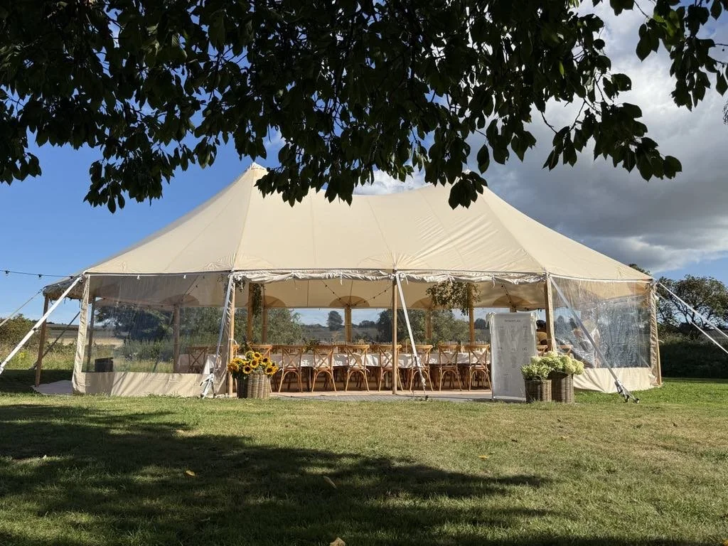 Et stort, hvidt, luksuriøst telt med åbne sider, opstillet af et professionelt team af eventpersonale, med store blomsterbuketter i flettede kurve og et velkomstskilt af stof ved indgangen.