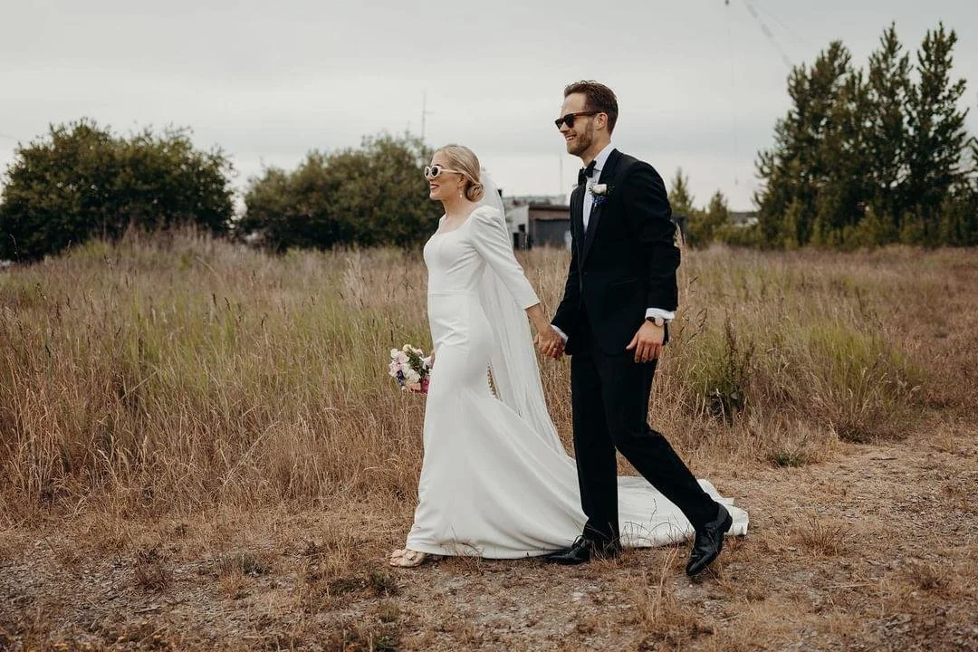 Bliss Cph - Et bryllup par holder i hånden og går gennem en rustik mark med højt græs, iført formelle outfits med brudekjole og smoking.