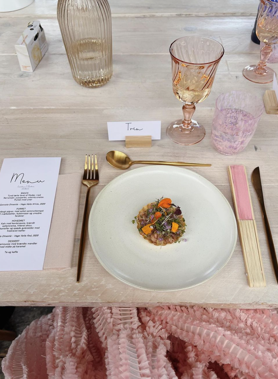 En borddækning med et fad med en lille anretning, en menu og bestik. Der er et glas hvidvin, et pink glas vand og en vandflaske. En pladsannonce med navnet 'Trine'.