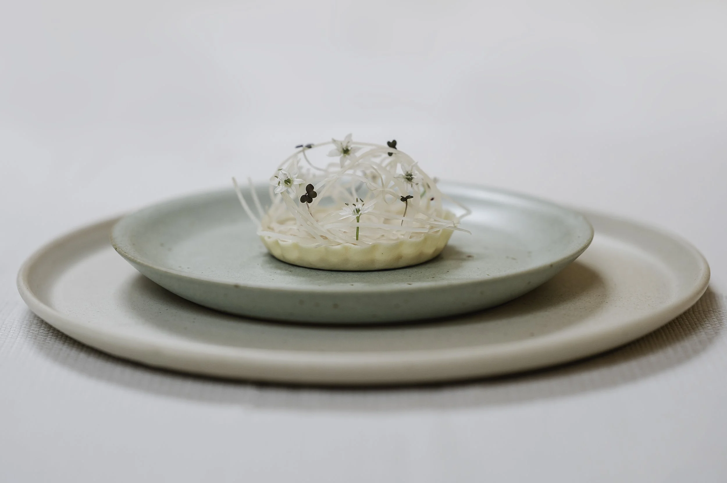 En skål med hvide blomster og små blade, placeret på to lag af flade tallerkener i pastelgrøn og cremefarvet.
