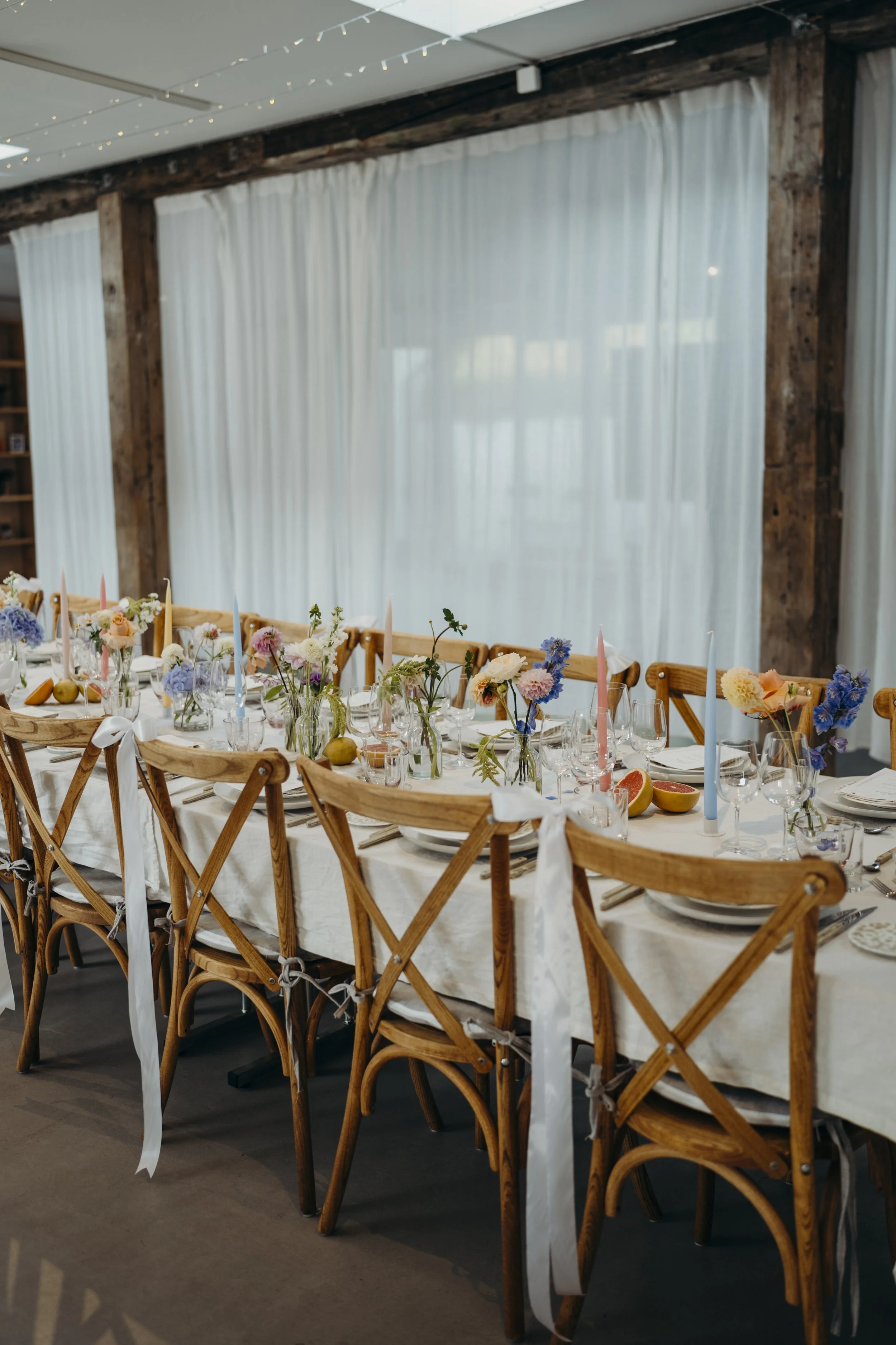 Lang bord med borddækning til en fest, pyntet med blomster, stearinlys, skåle med frugt og bestik.