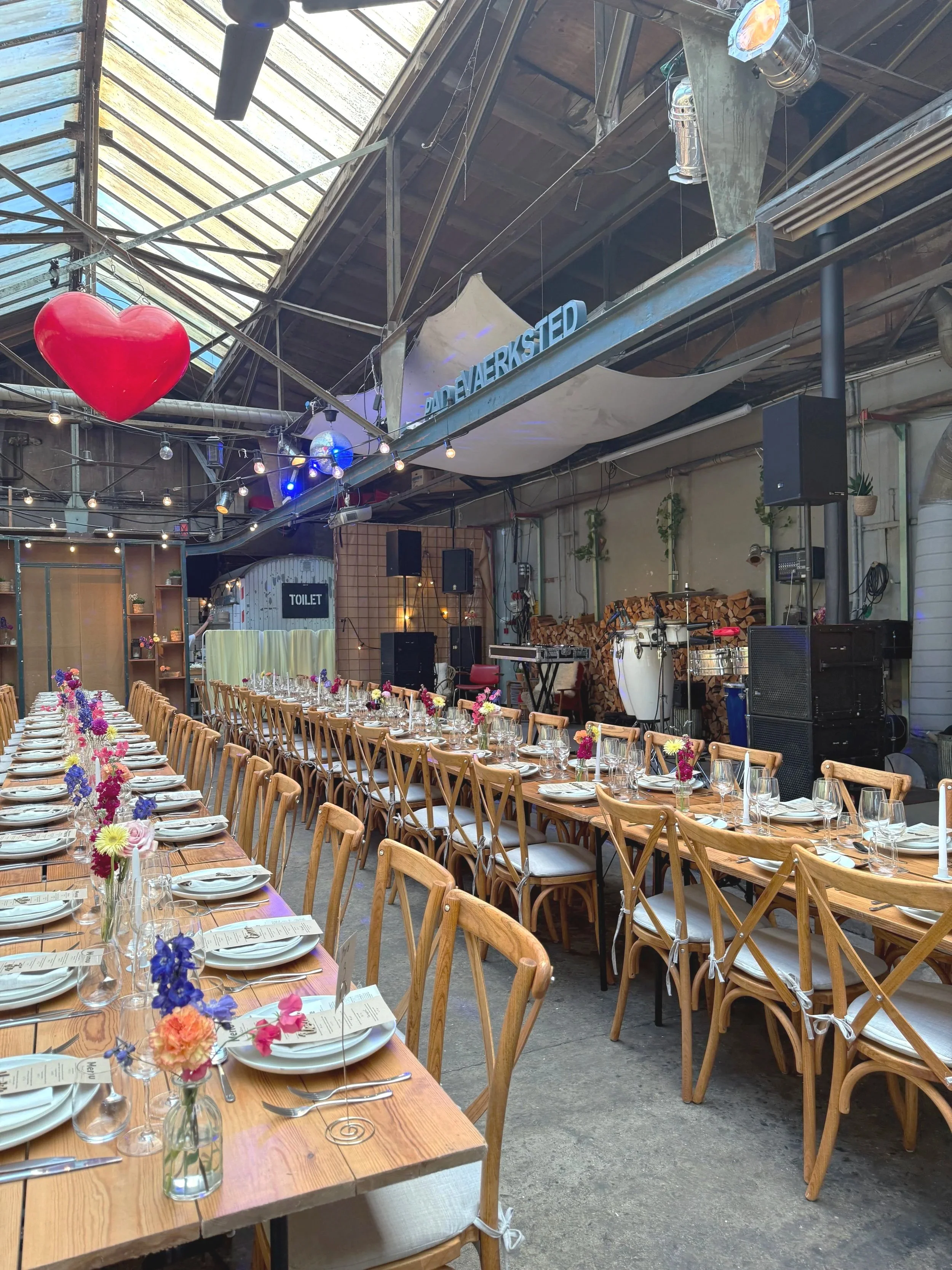 Rå industrielt bryllupssted i de gamle skibshaller med lang borddækning, blomsterdekorationer, pyntede glas og tallerkner, og en scene med musikudstyr. Der hænger en stor rød hjerteballon og et skilt med teksten 'Pladeværkstedet'.