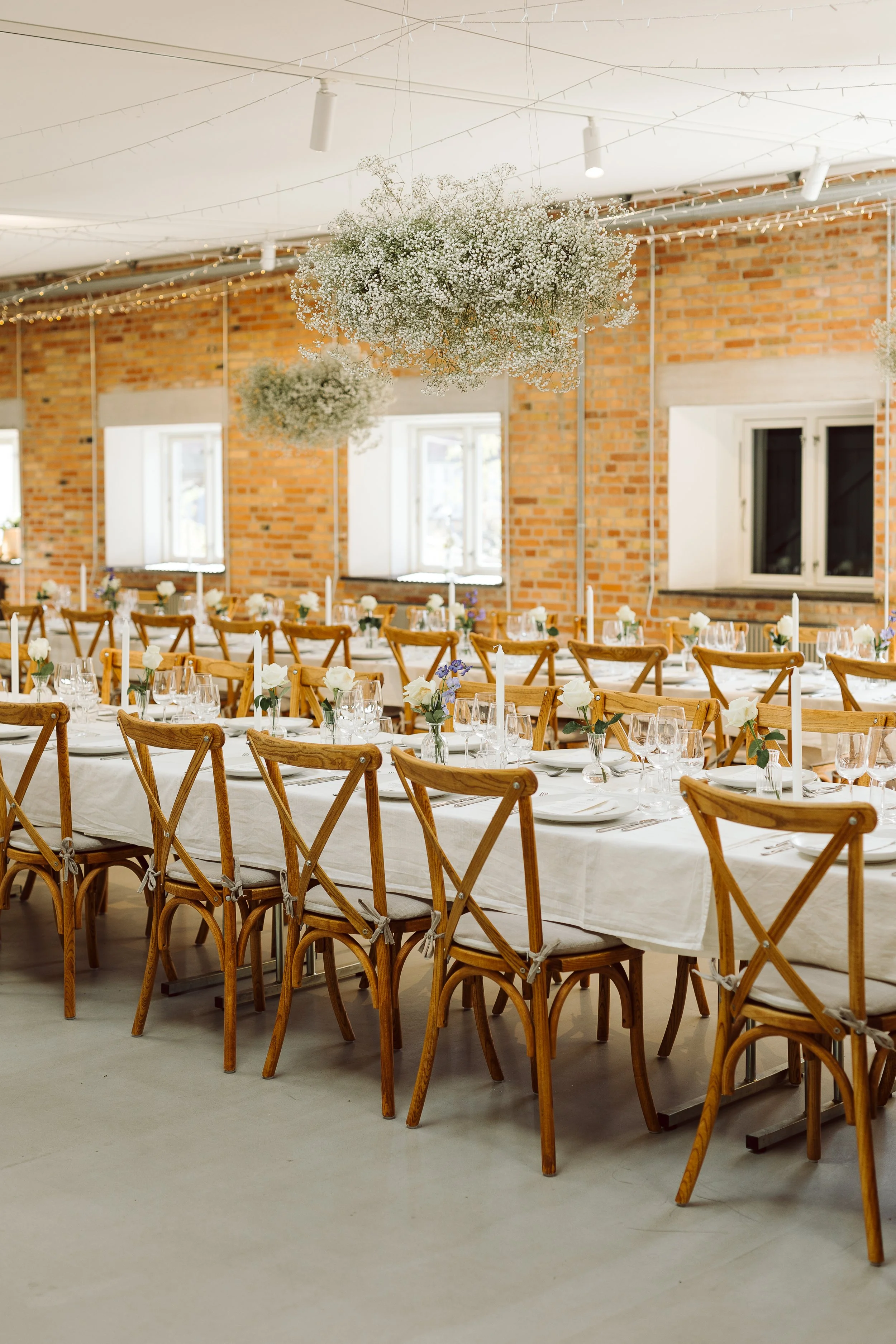 Indretning til fest med lang plads buffet, hvide andeglas, blomster i vaser, lysestager og hvide servietter, hængende blomsterdekorationer og loftslys.
