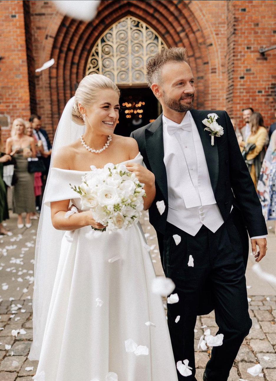 Et brudepar går ud fra kirke, omgivet af gæster, med blomsterknas i luften.