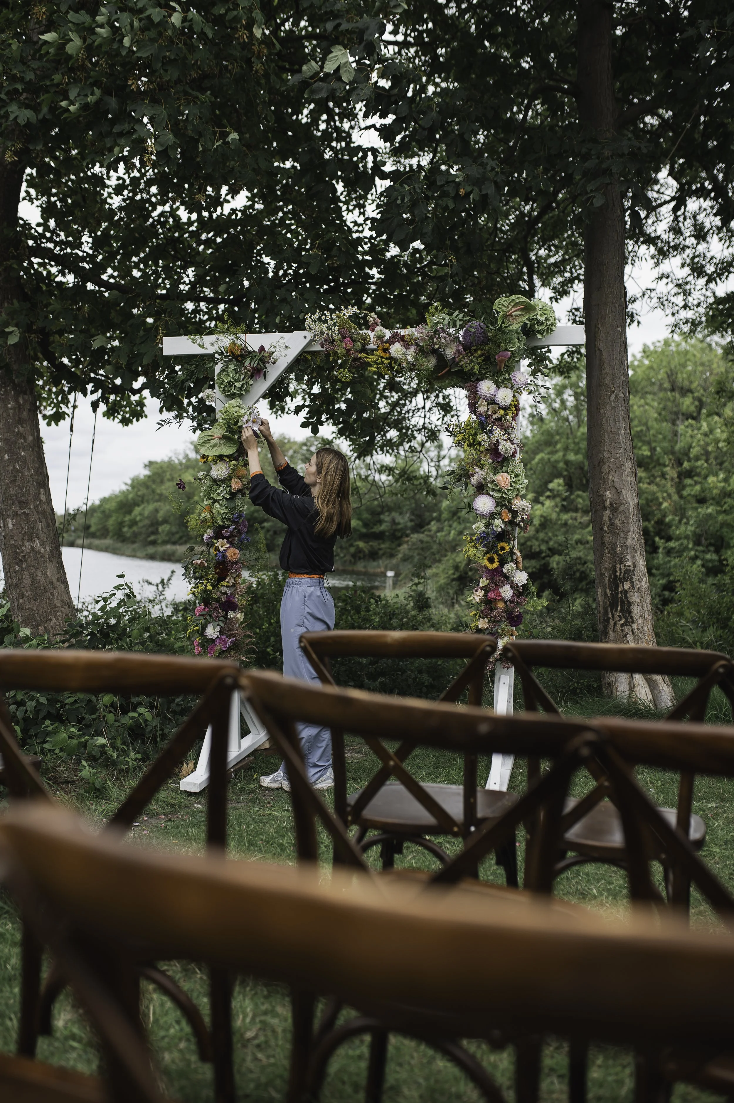 Bliss CPH – En blomsterdekoratør fra Holly Flowers hænger blomster på bryllup bue foran en sø i en grøn park, klar til en udendørs ceremoni.