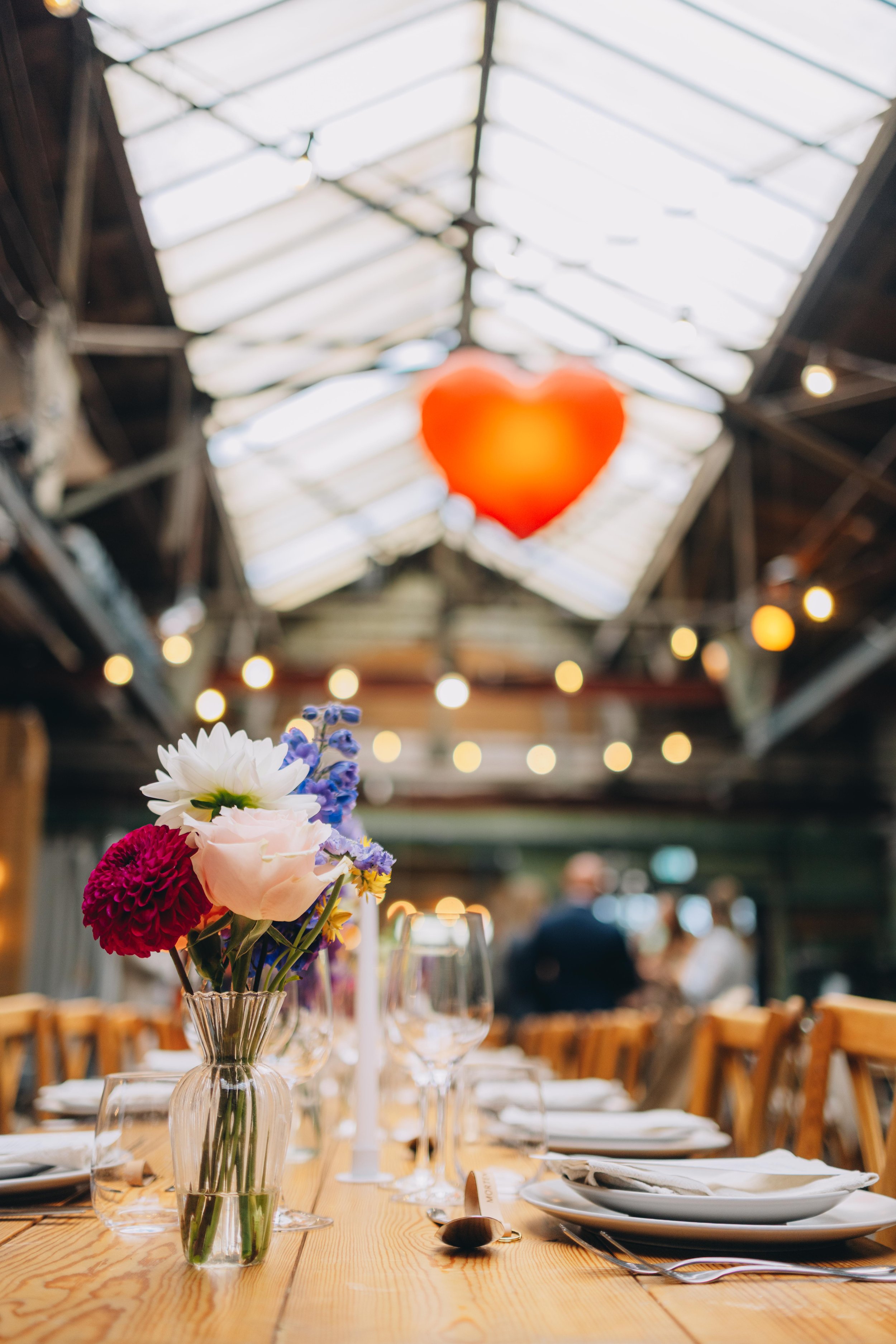 Bliss CPH – En lang træbord med blomsterdekorationer, sat til en fest i en rustik bygning med højt glasloft og udsmykket