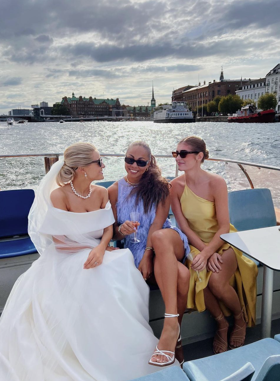 Tre kvinder sidder på en båd, iført dress, og nyder en dag på vandet med en by i baggrunden.