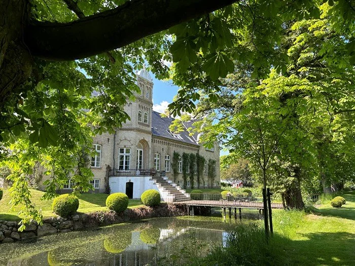 Bliss Cph - Søllestedgård Gods på Lolland, en slot i en grøn have med en dam foran, omgivet af træer og buske, under en klar blå himmel.