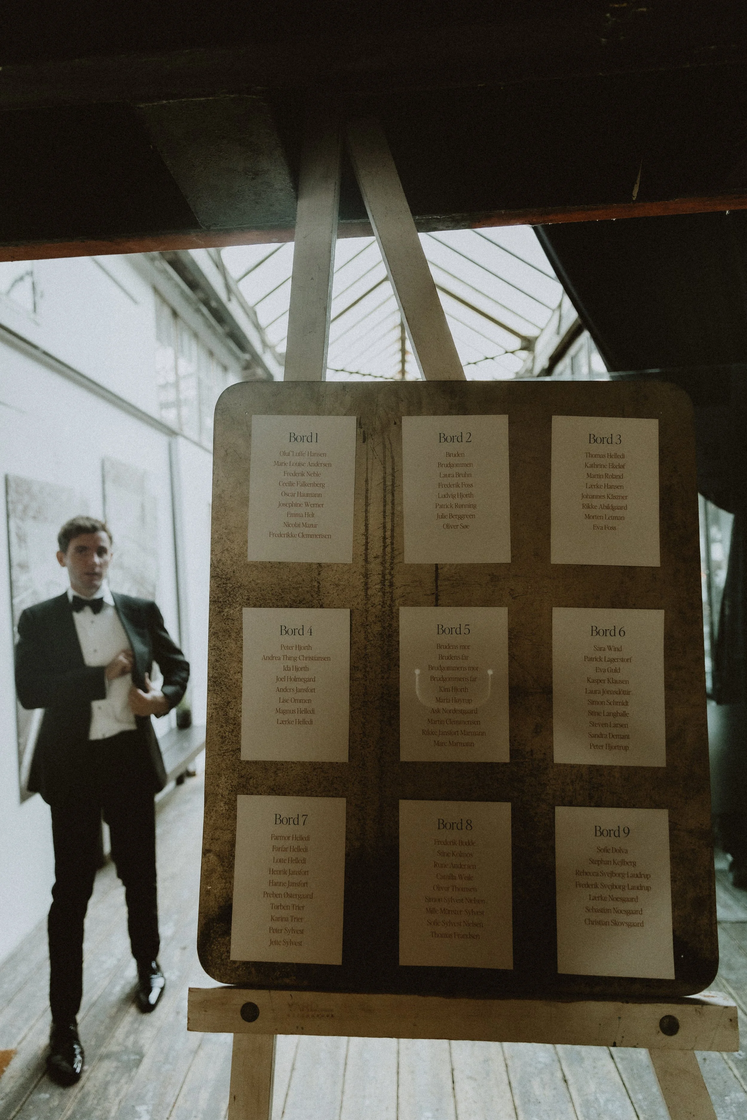 Bryllupsbordplan på en plade af rå metal og et kunstnerstaffeli, med en mand i sort smoking i baggrunden.