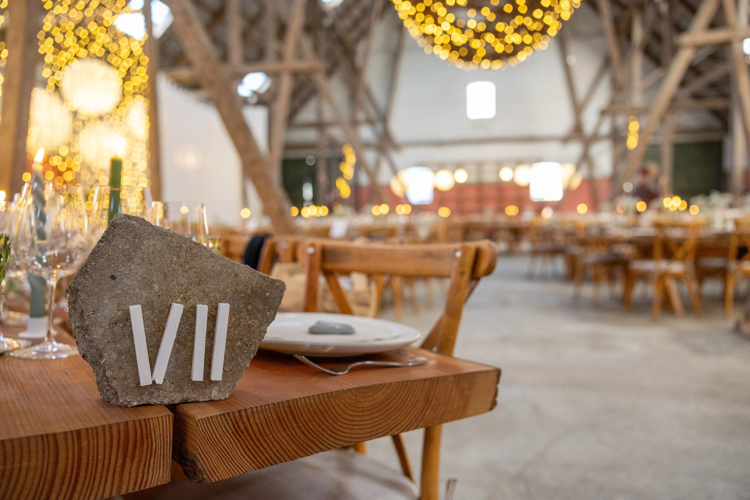Et festligt bord med 7 lignende glas, sat til en fejring i en stor, rustik hal med træbjælker og julestjerner, og et stenpynt med roman numeral VII på bordet.