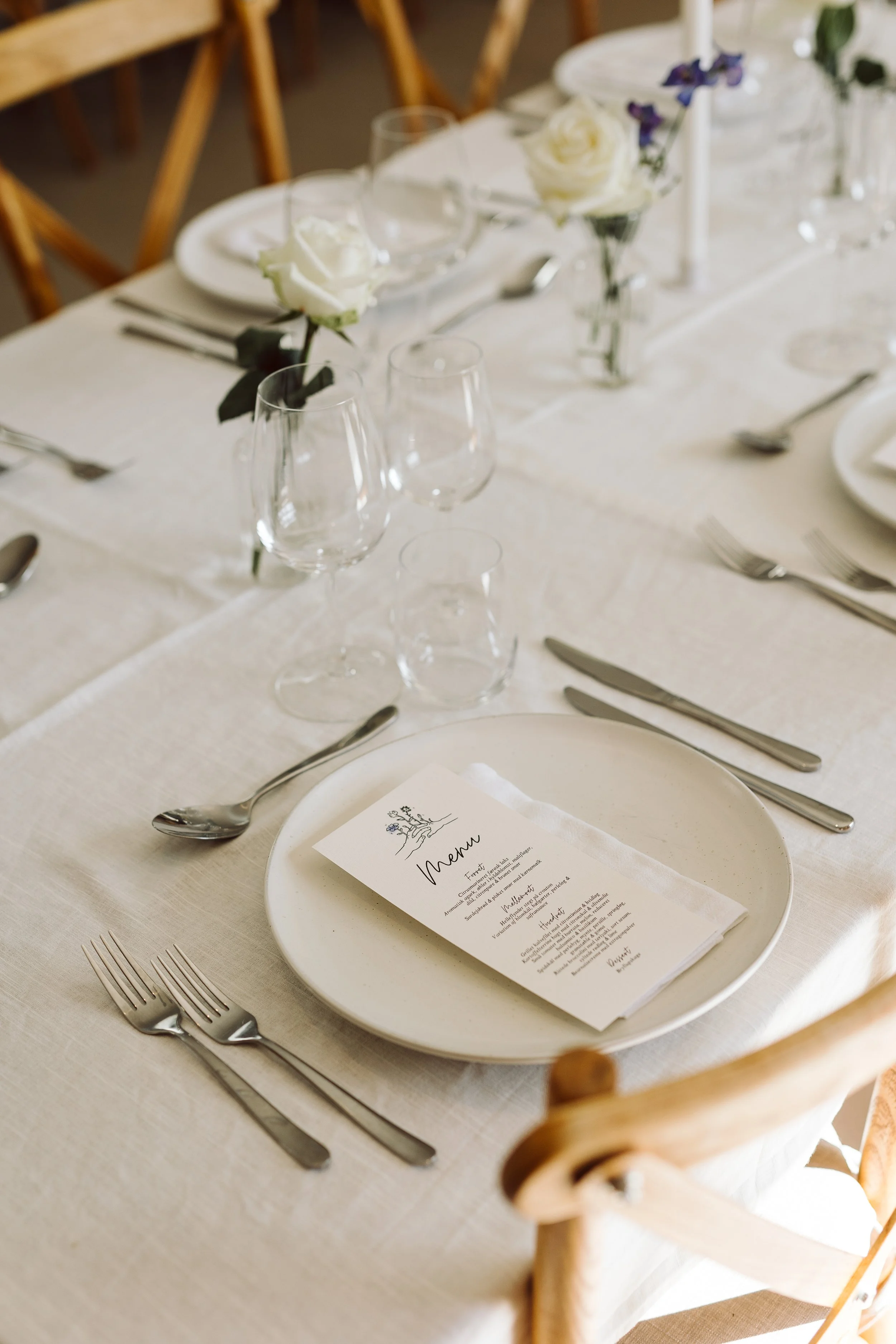 Elegant bryllupstafel med hvide tallerkener, sølvbestik, vin- og vandglas, blomsterdekoration og menu på tallerkenen.