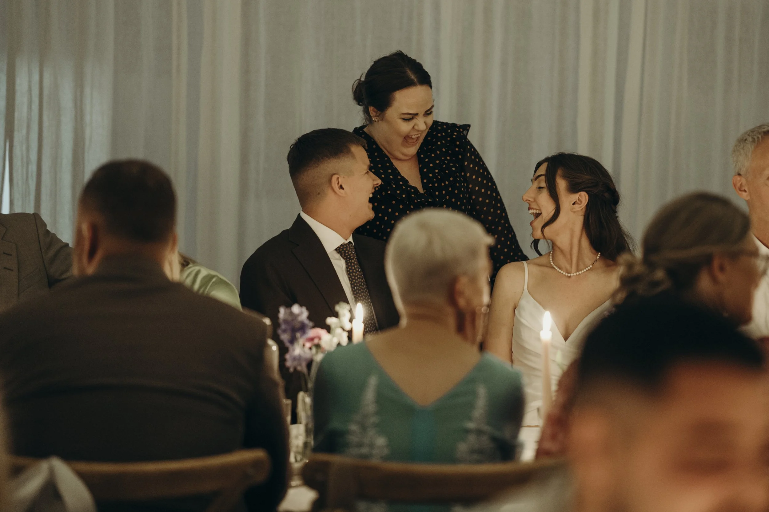 Glade mennesker til en fejring, hvor en kvinde giver en gave til en mand og en kvinde, omgivet af andre gæster.