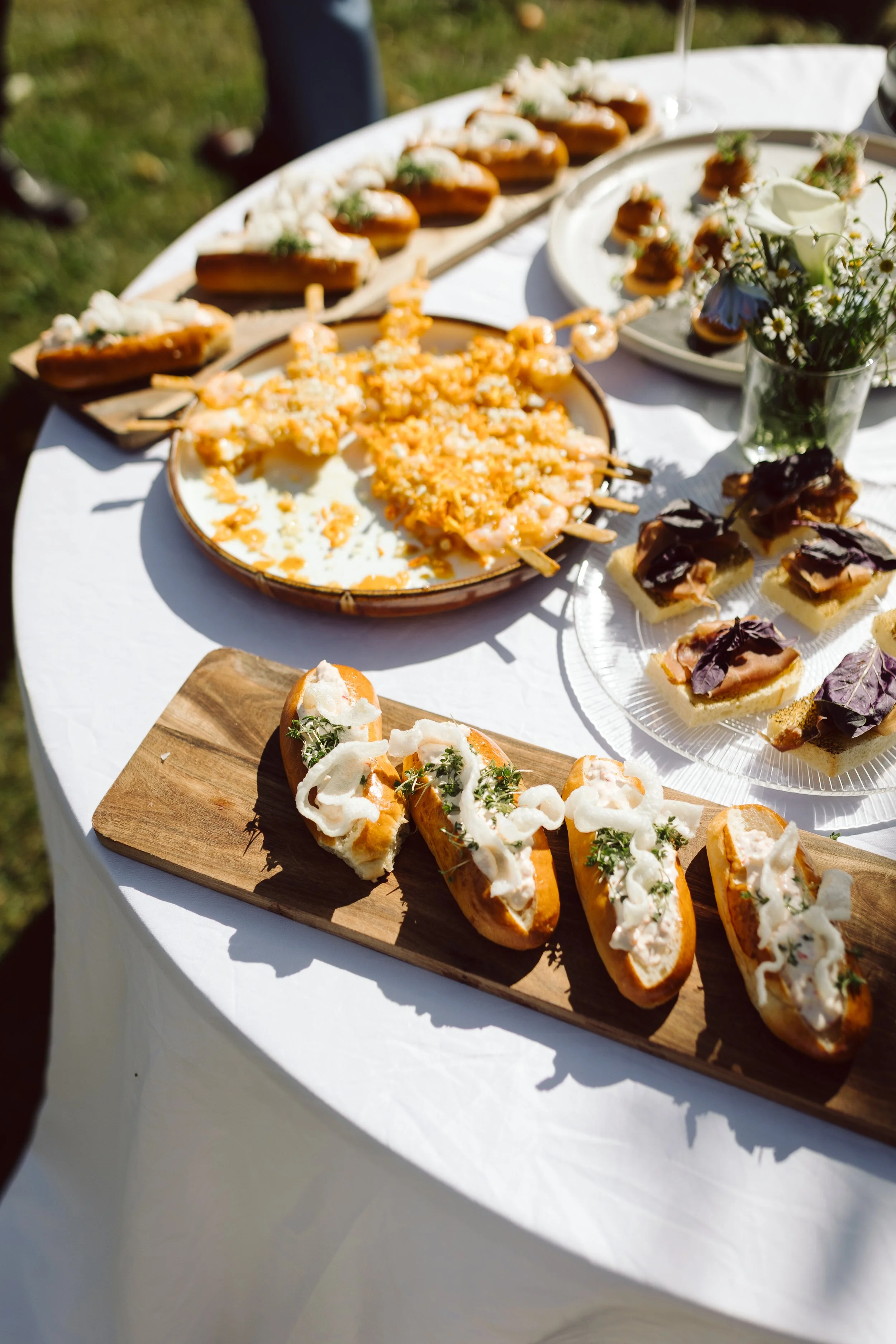Udvalg af småretter og fingerfoods på et hvidt dækket bord, inklusive små hotdogs med fyld, skåle med skaldyrsspyd, små sandwicher med kød og grøntsager, samt en vase med blomster.