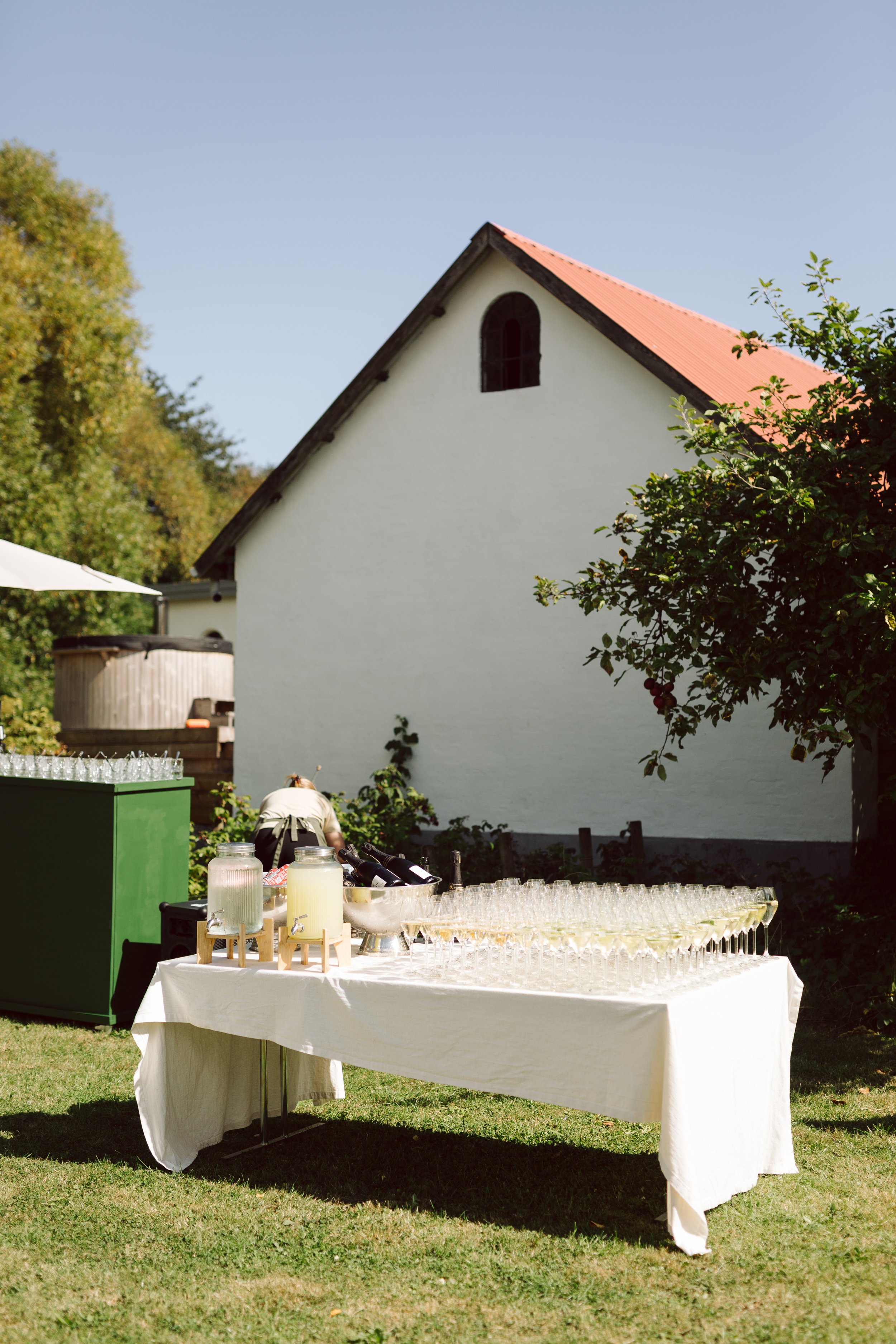 Udendørs drikkebord med glas og store termoser foran en hvid bygning med rødt tag, omgivet af grønt tilbehør og planter.