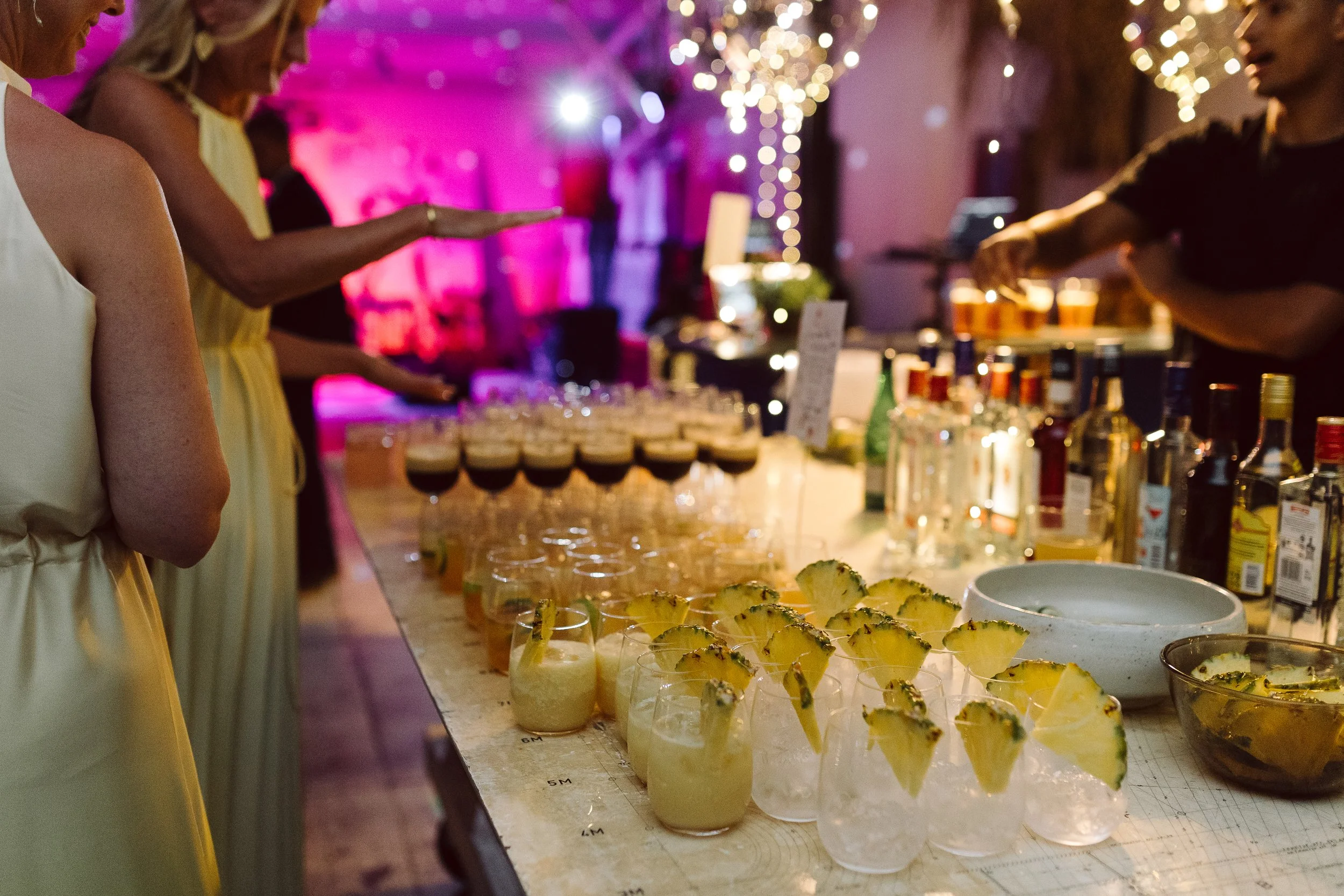 Billedet viser en bar med forskellige drinks og cocktails, folk der danser, og festlig belysning med lysende dekorationer.