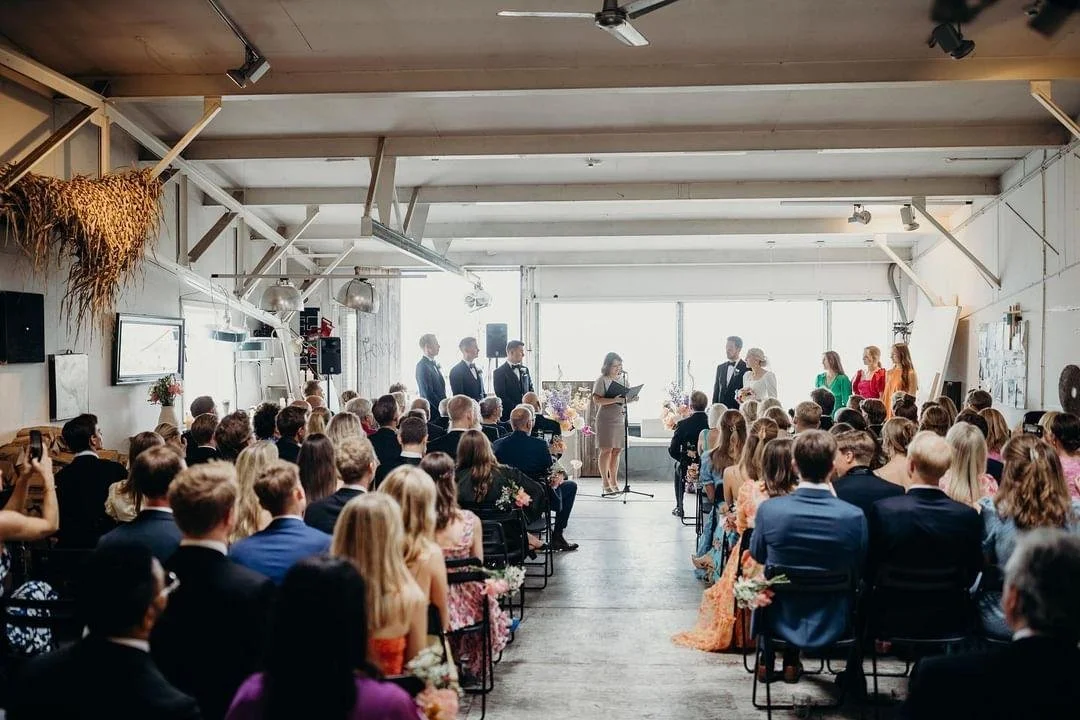 Bryllupsceremoni med brudepar og forlover foran en stor forsamling i et lyst industrilokale.