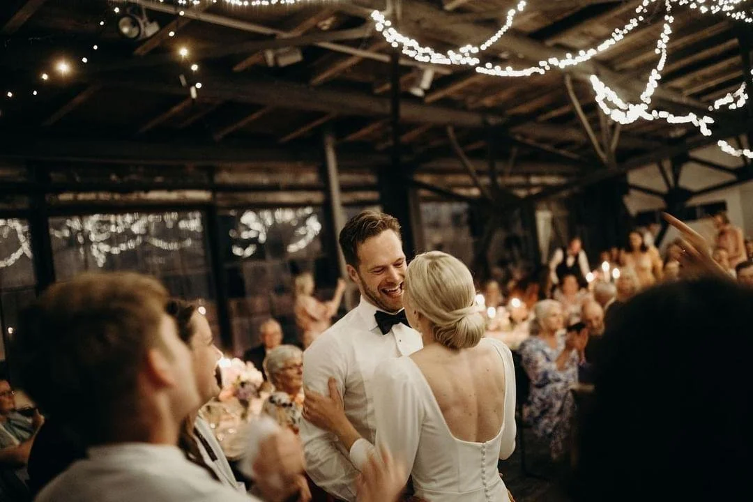 En mand og en kvinde danser romantisk på en festlig aften, omgivet af familie og venner, med pyntede lys overalt.