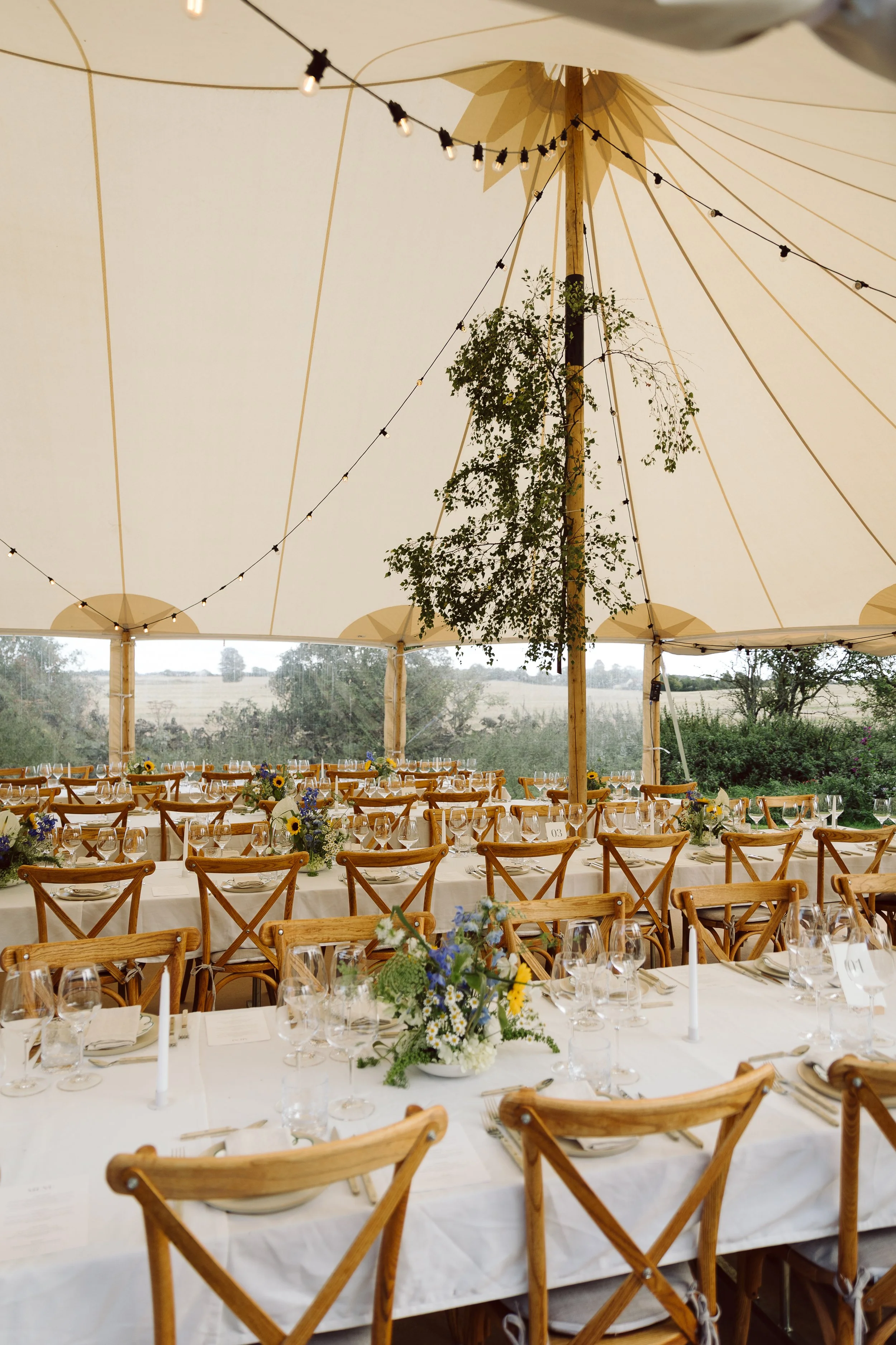 Indendørs festtelt med langbord dækket med hvide duge, blomsterdekorationer, glas og bestik, træstole, og lyst luftlys under et blødt stofloft med trækonstruktion og grønne planter.