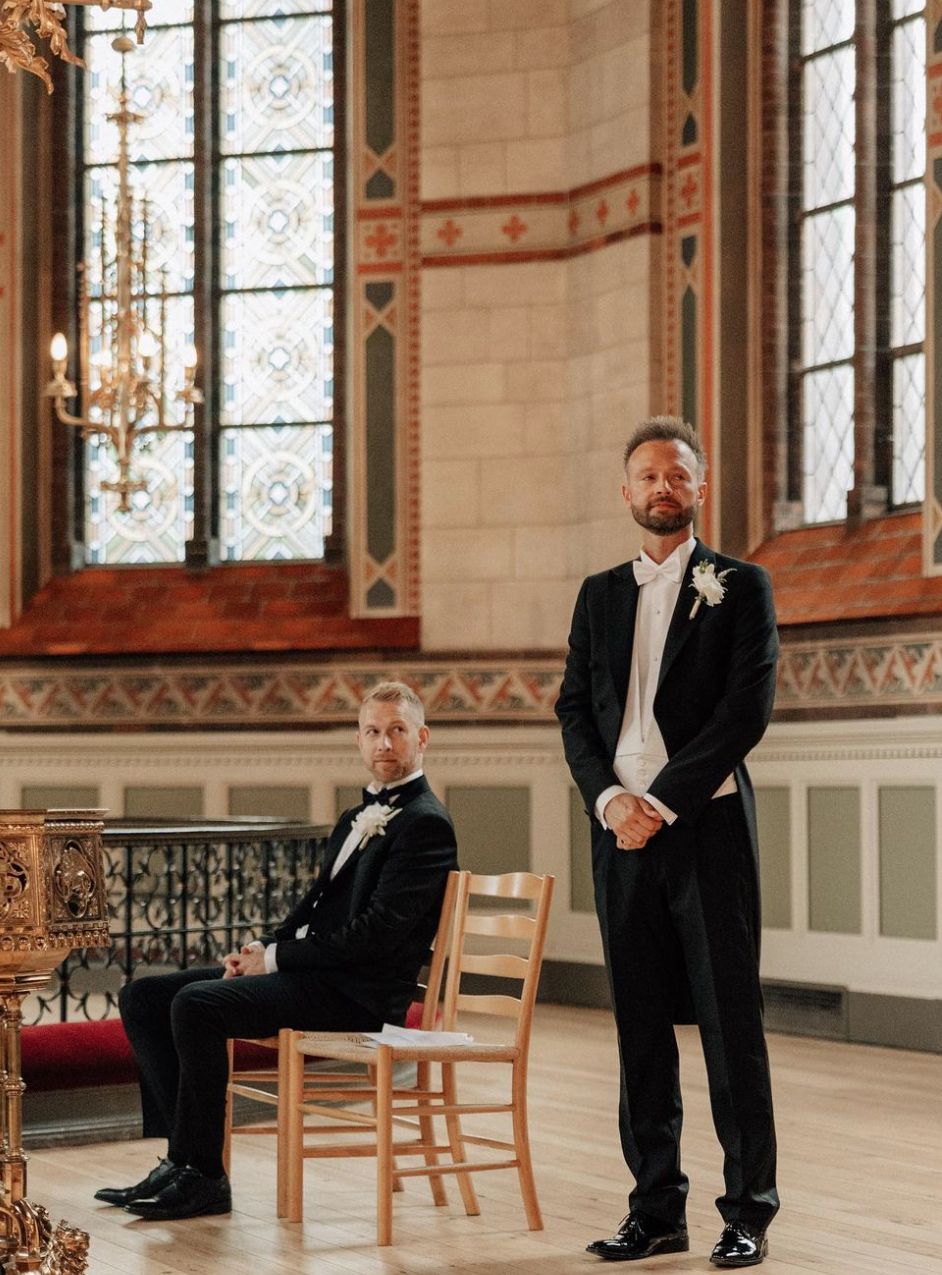 To mænd i formelle sort-tweed tøj, den ene sidder på en træstol og den anden står, i en smuk historisk kirke med farverige vægge og store farvede vinduer