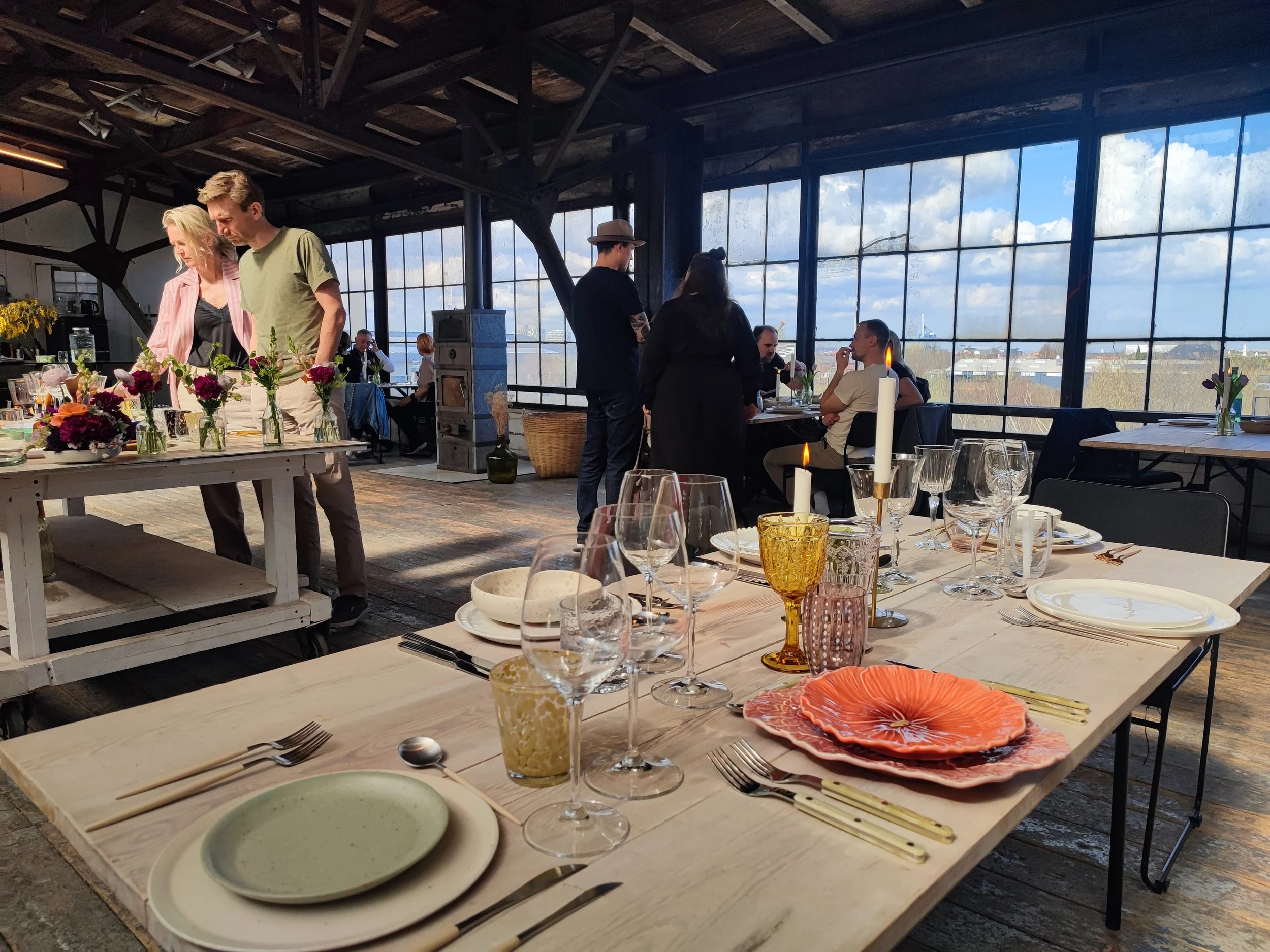 Indendørs restaurant med borddækning, stearinlys, duer, gæster og store vinduer med udsigt til blå himmel og skyer.