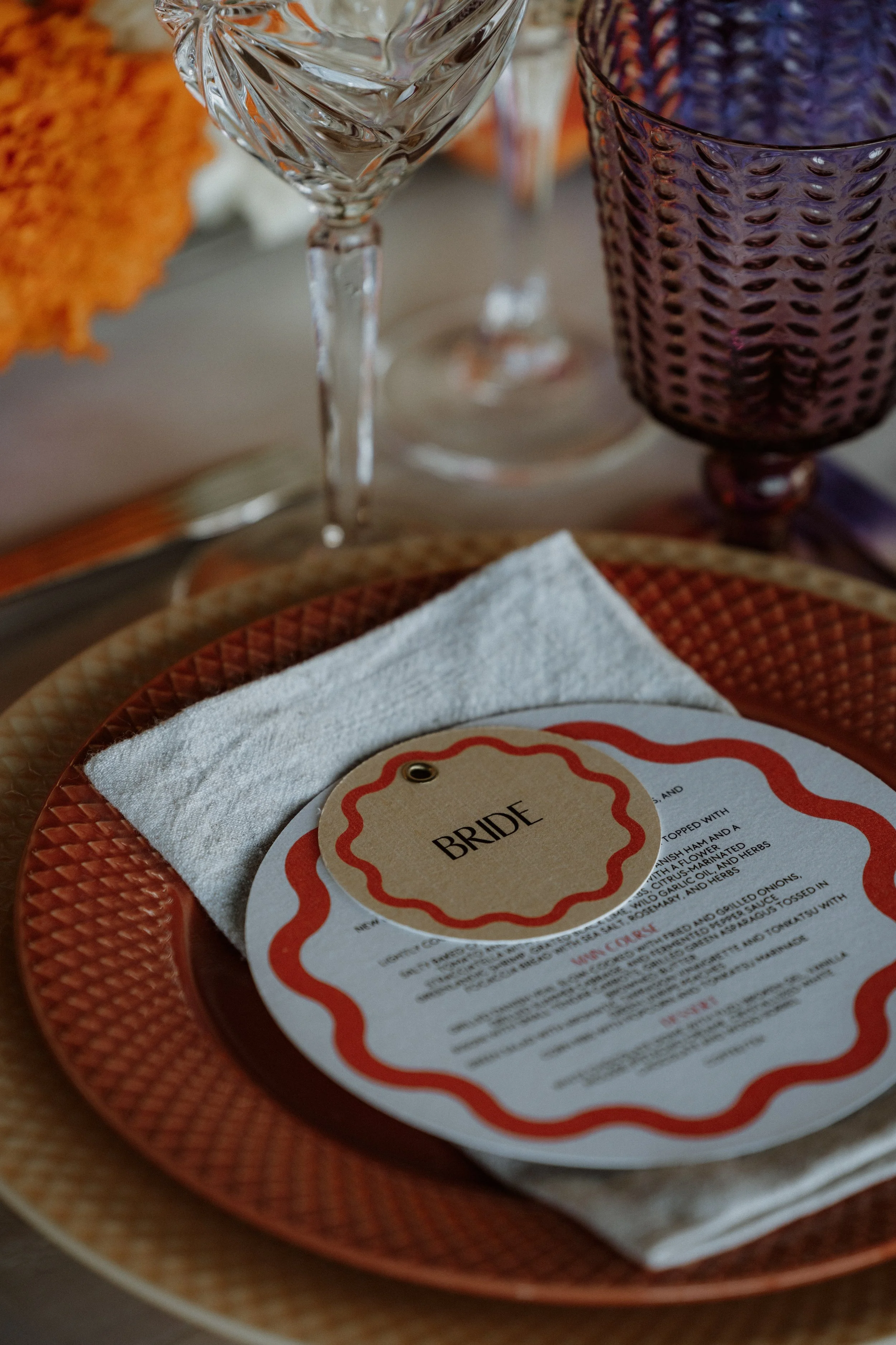 En festlig borddækning med en tallerken, en knap med teksten 'BRIDE', en hvid serviet, to glas og en mulepose i baggrunden.