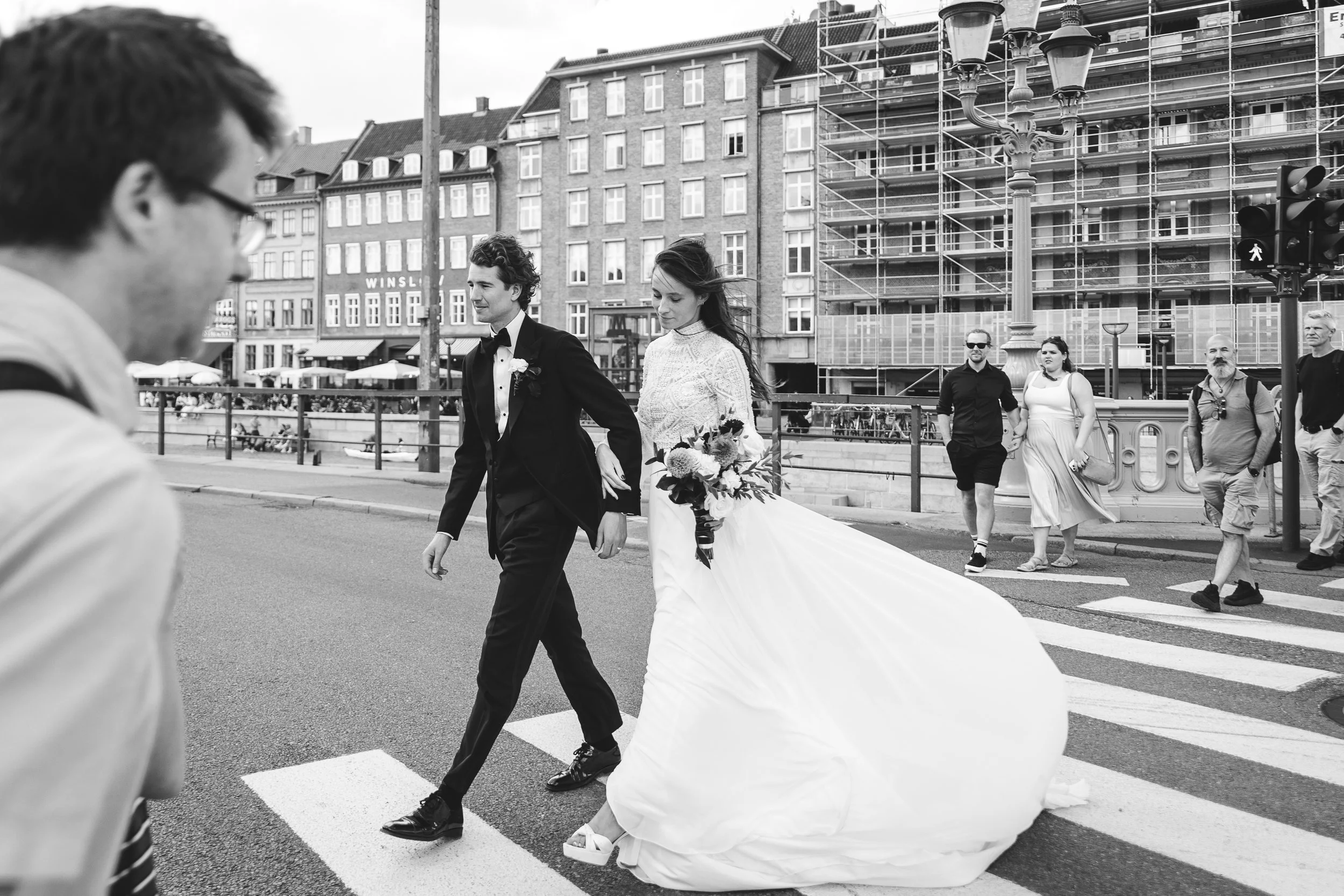 En brud i lang kjole med lyksalighedens blomster i hænderne krydser gaden, ledsaget af sin mand i sort smoking. Der er fodgængere og gamle bygninger i baggrunden, når de krydser gaden i Gammel Strand, København.