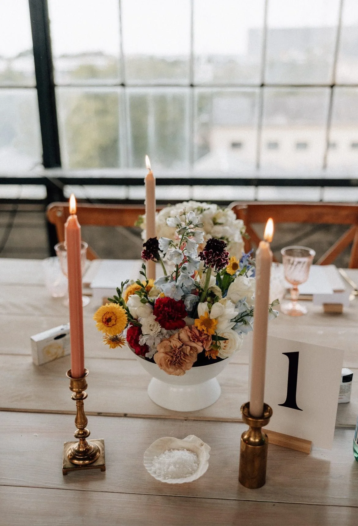 Borddekoration med lysestager, blomsterdekoration og bordnummer "1" på en træbord, udsigt gennem vindue i baggrunden.