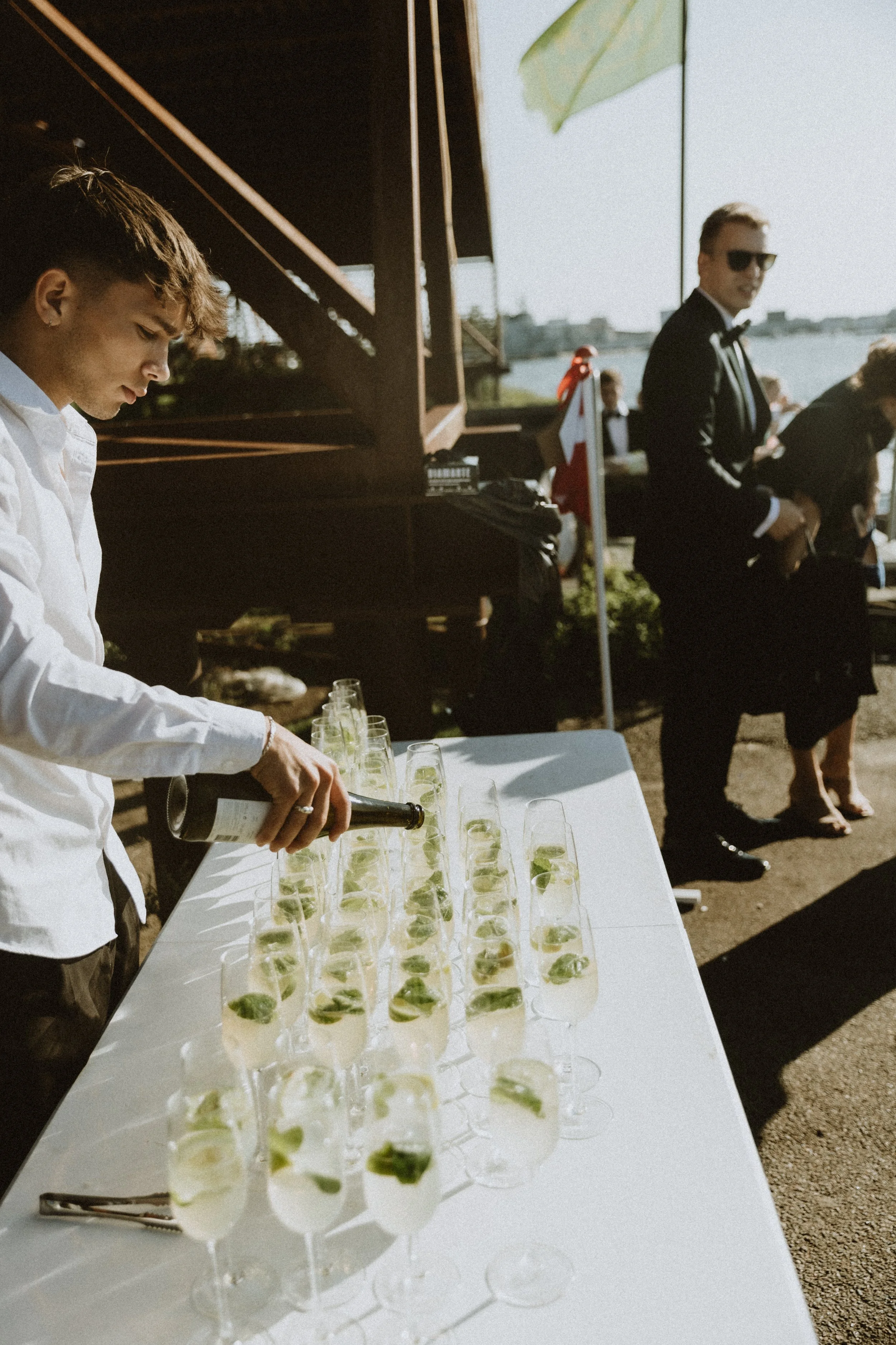Bliss CPH – En professionel tjener, der hælder champagne i champagneglas med lime, ved en udendørs festivitet ved vandet, med andre gæster