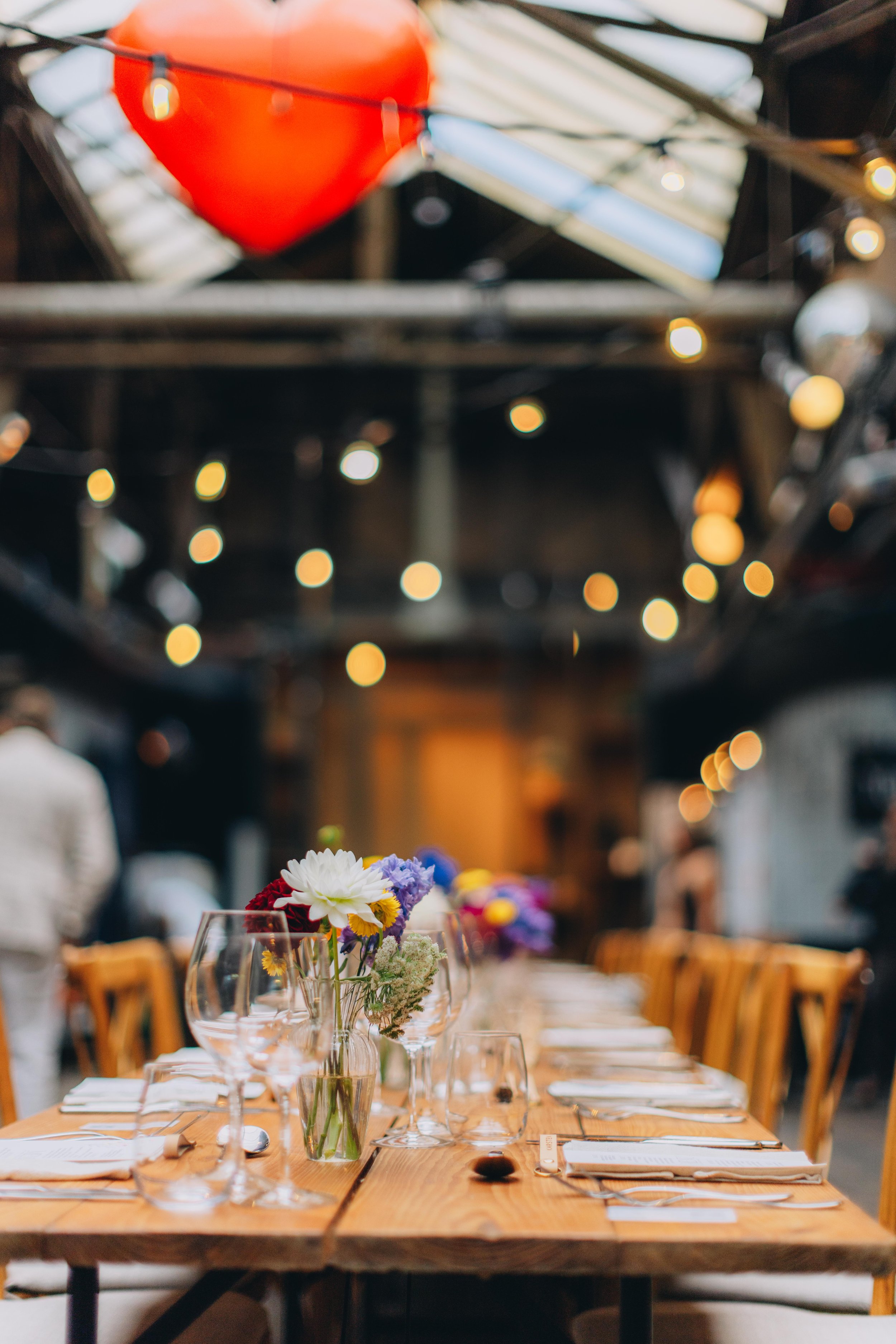 Lang bord med blomster i vaser, sat op til et måltid, i et festlokale med bløde belysning og blødt belysning i loftet.