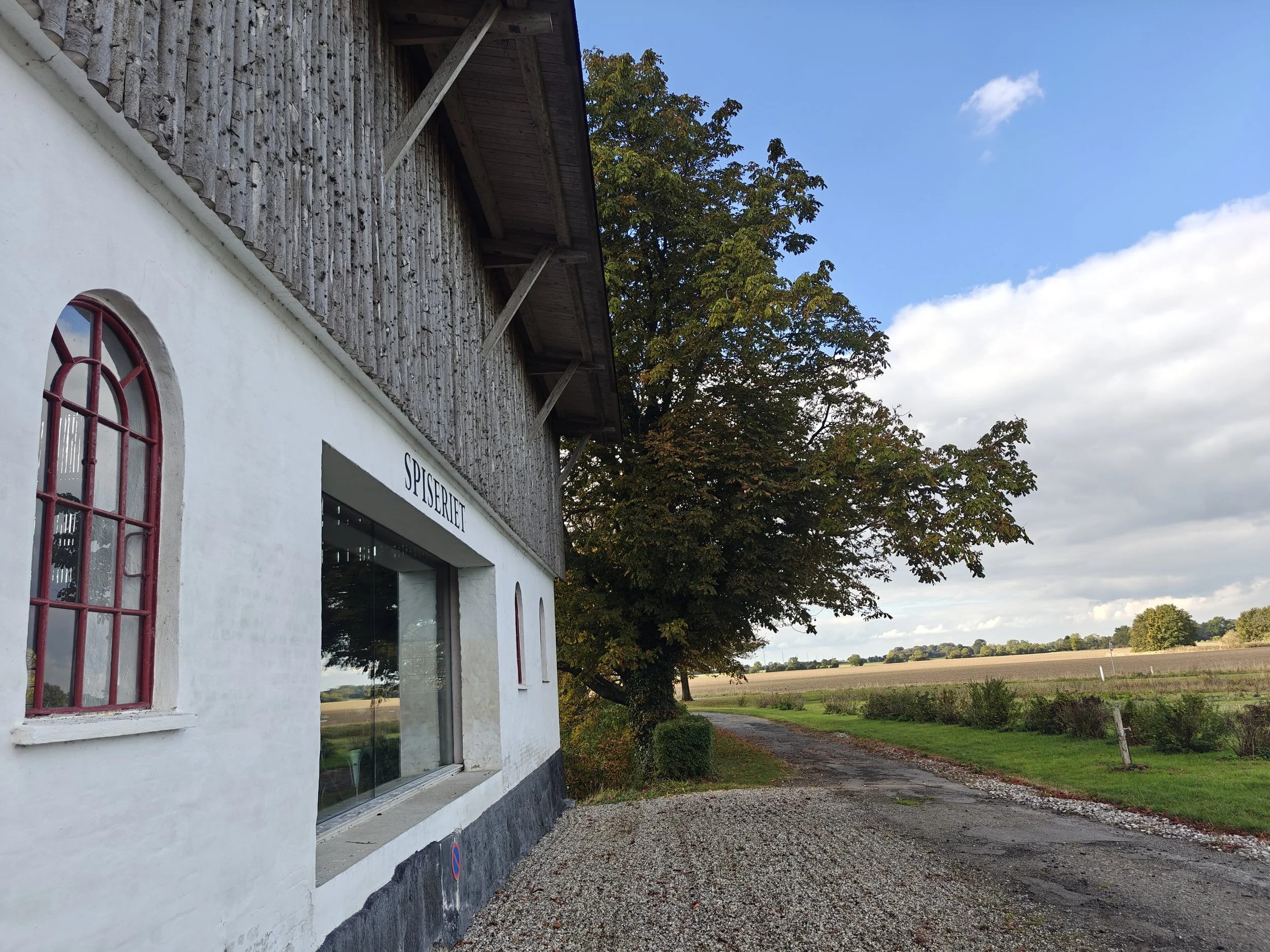 Bliss Cph - Moderne, dansk arkitekttegnet, hvid bygning med et stort vindue, der vender ud mod marker, afgrøder og et stort træ.