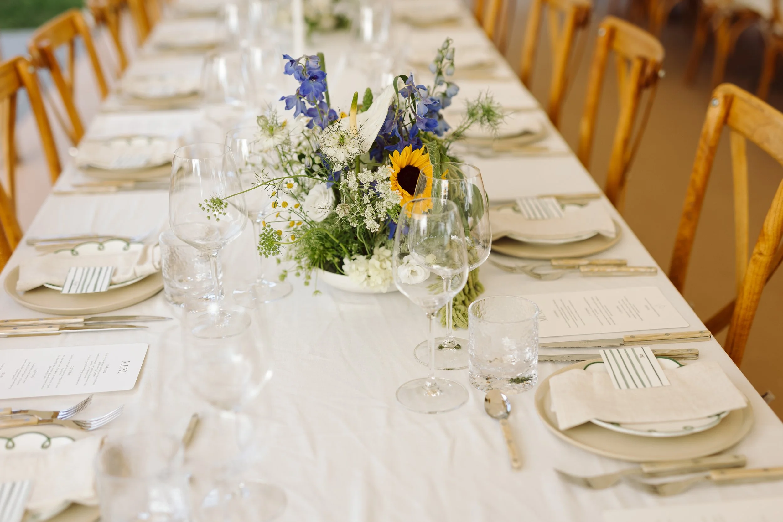 Borddækning med blomstercenter, vin- og vandglas, tallerkener, bestik og menusedler, klar til fest.