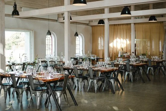Bliss Cph - Søllestedgård event venue med flere borde dækket til middag og vin, moderne industriel lysarmaturer og store vinduer.