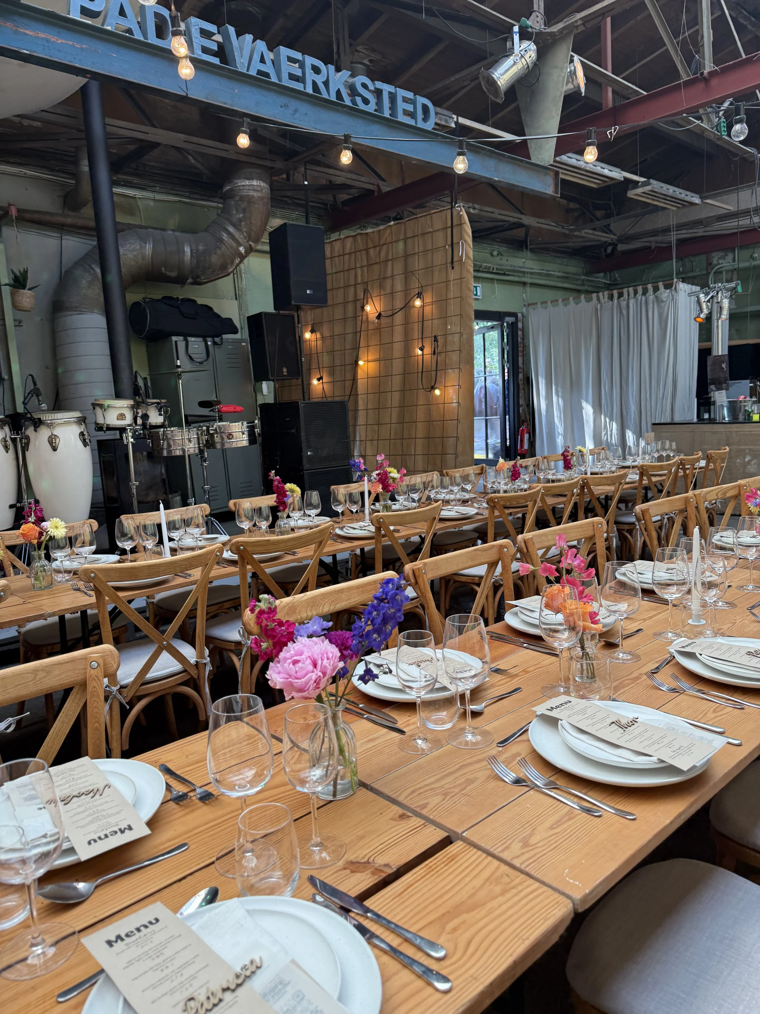 Bliss CPH – Rå festlokale med langborde dækket med tallerkener, glas og bestik. Der er blomsterdekorationer og en stor scen