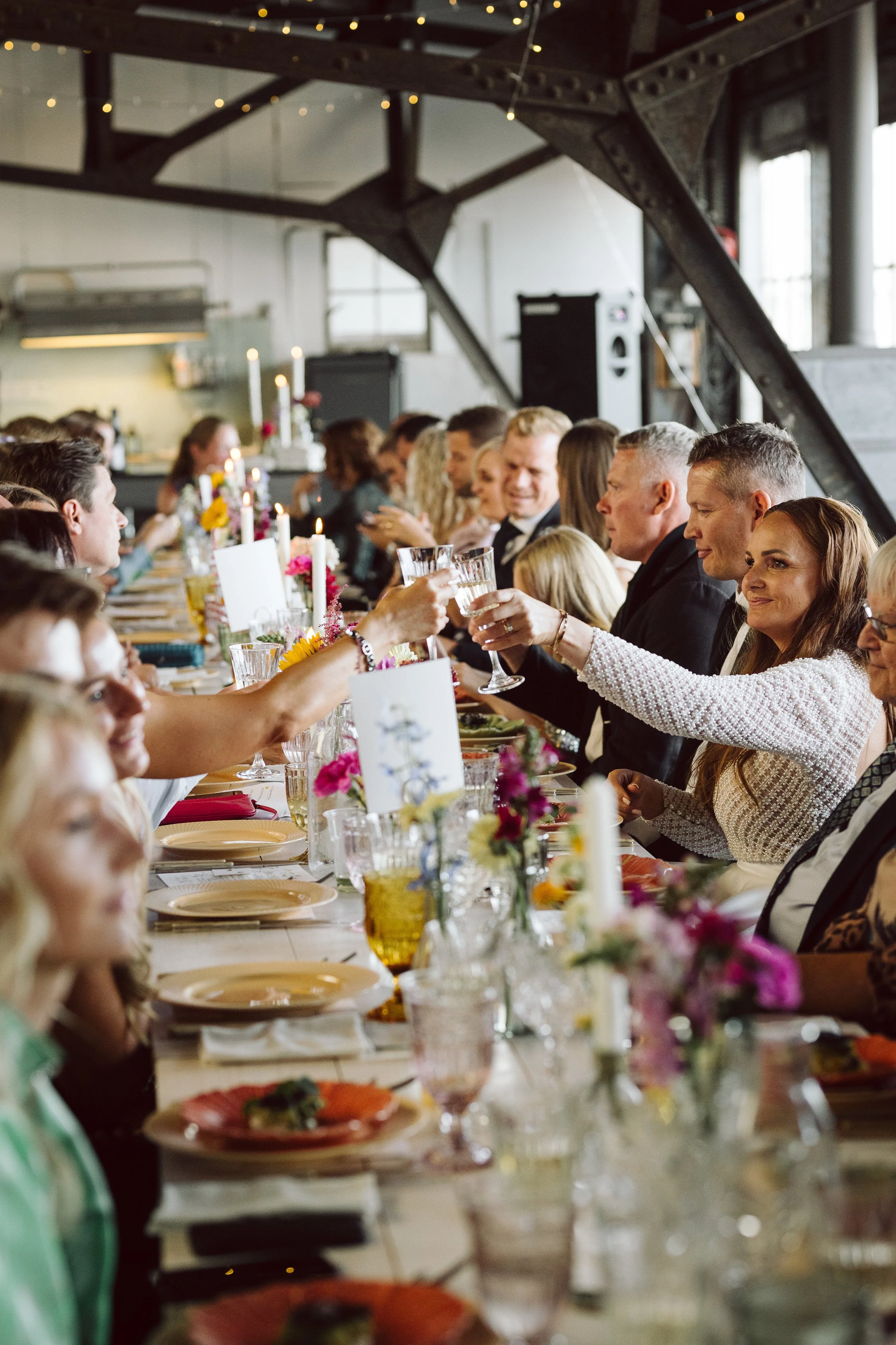 Folk samles omkring langbord til fest, deler drinks, med blomsterdekorationer og stearinlys.