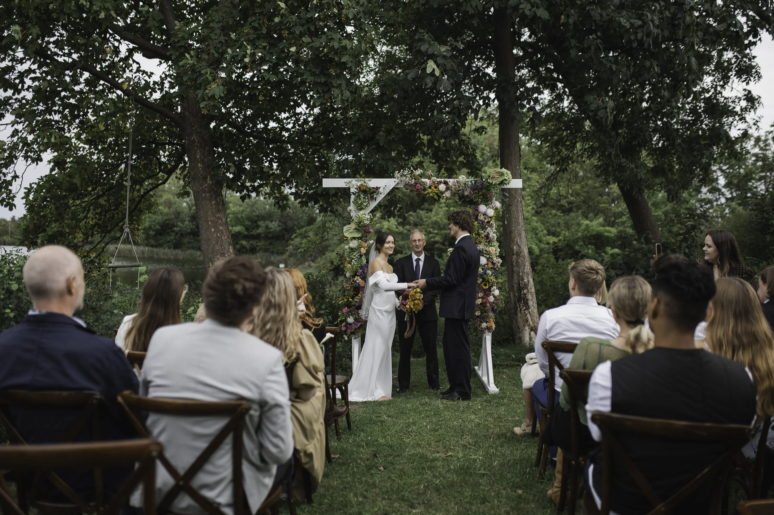 Et udendørs bryllup med en ceremoni foran et blomsterdekoreret ramme mellem to træer, hvor bruden og gommen står foran en dommer. Gæster sidder på træstole og observerer ceremonien, hvilket foregår ved en græsplæne ved en sø eller et vand.