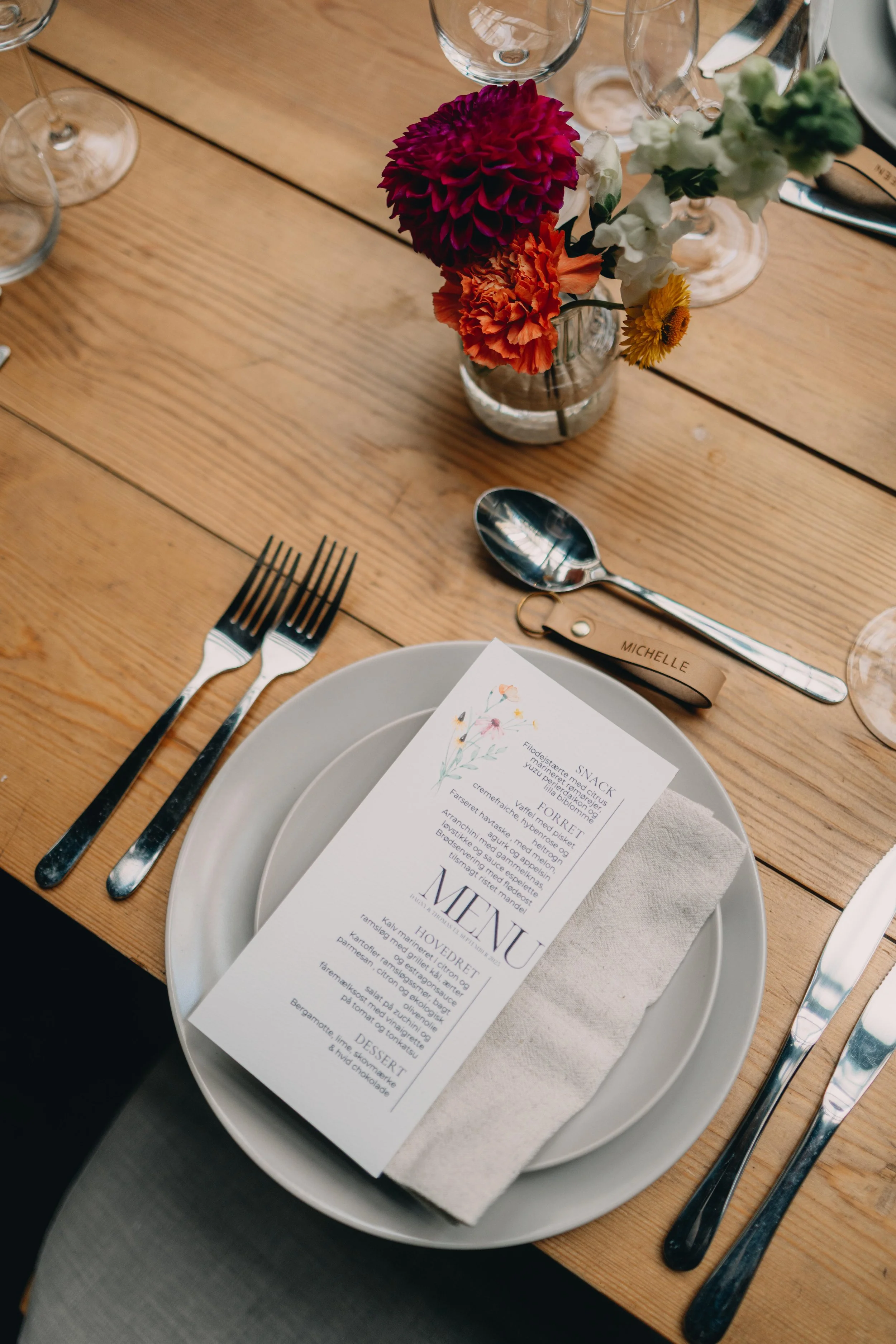 Bordsetning med tallerken, bestik, glas, vase med farverige blomster, menyark, og personlige navneskilte.