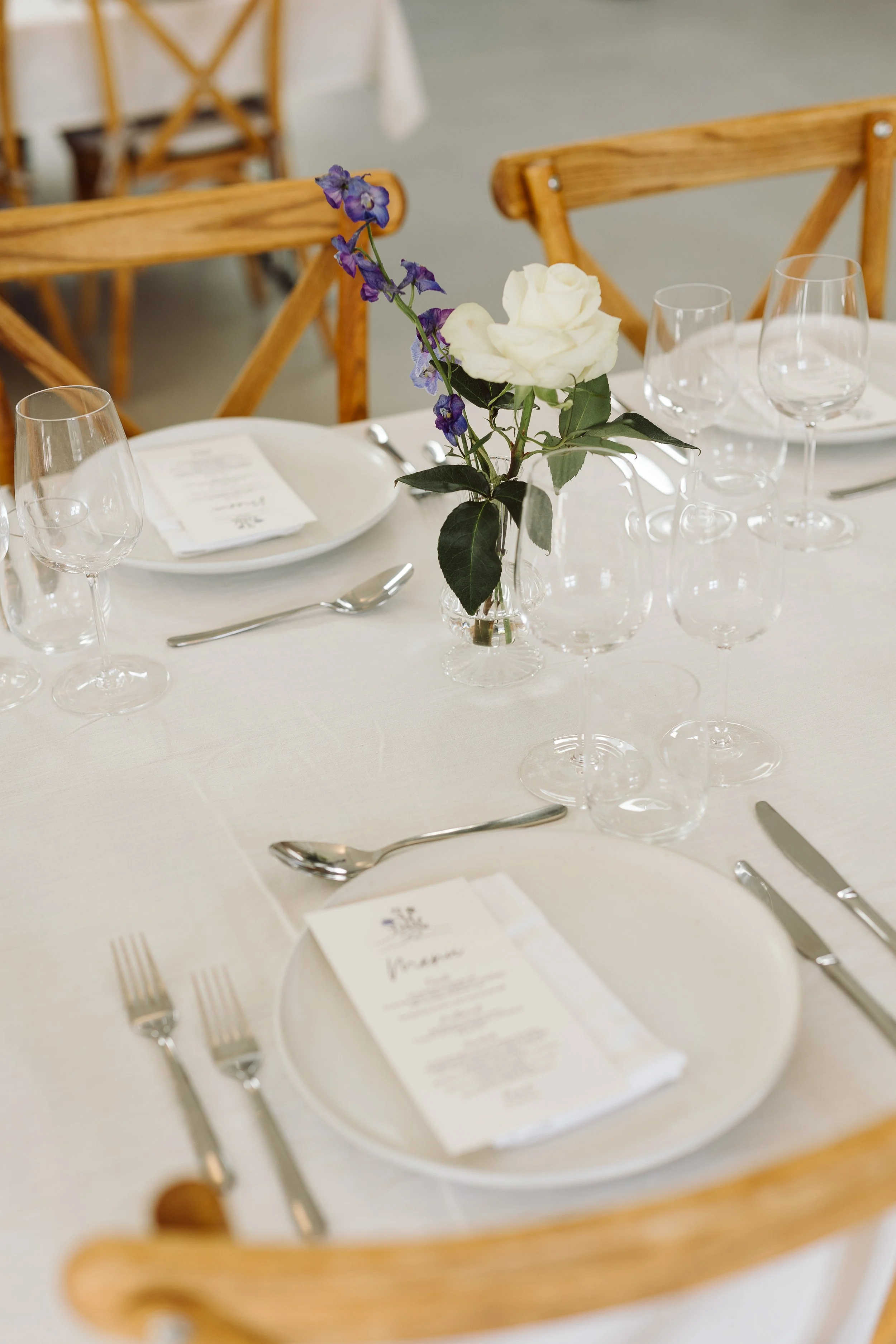 Et bord dækket til en fest eller reception med en hvid dug, glas, tallerkener, bestik og en lille vase med hvide og lilla blomster.